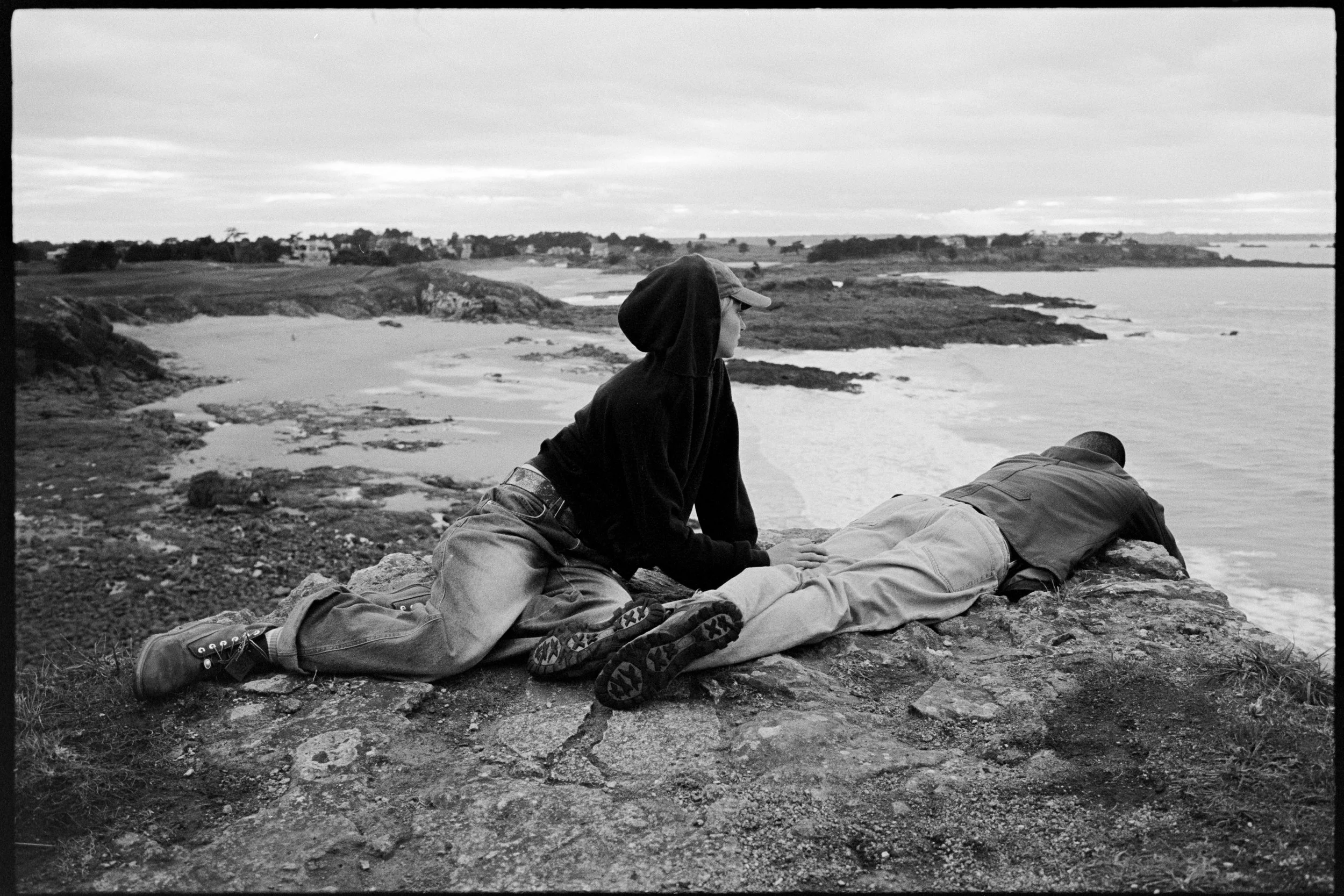 Deux personnes allongées sur un rocher au bord de la mer, l'une face à la plage et l'autre de dos, avec un paysage côtier en arrière-plan.