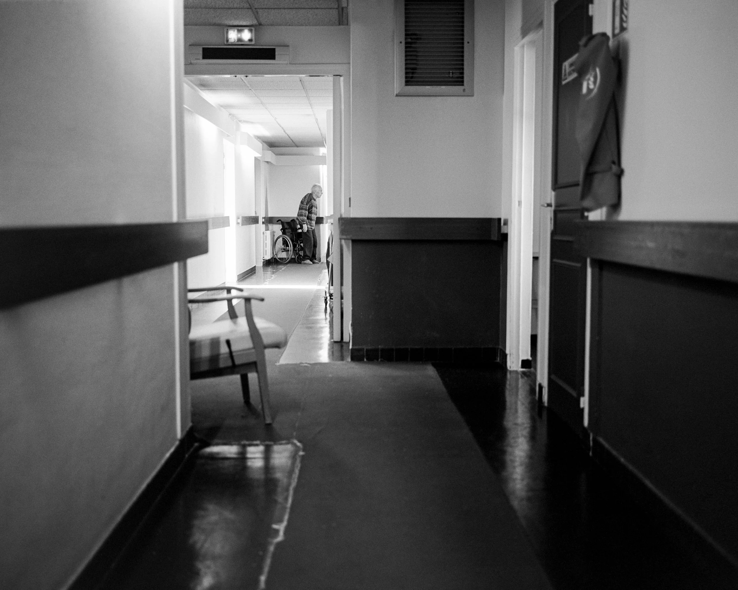 Un couloir dans un établissement de soins avec une personne en fauteuil roulant au fond.