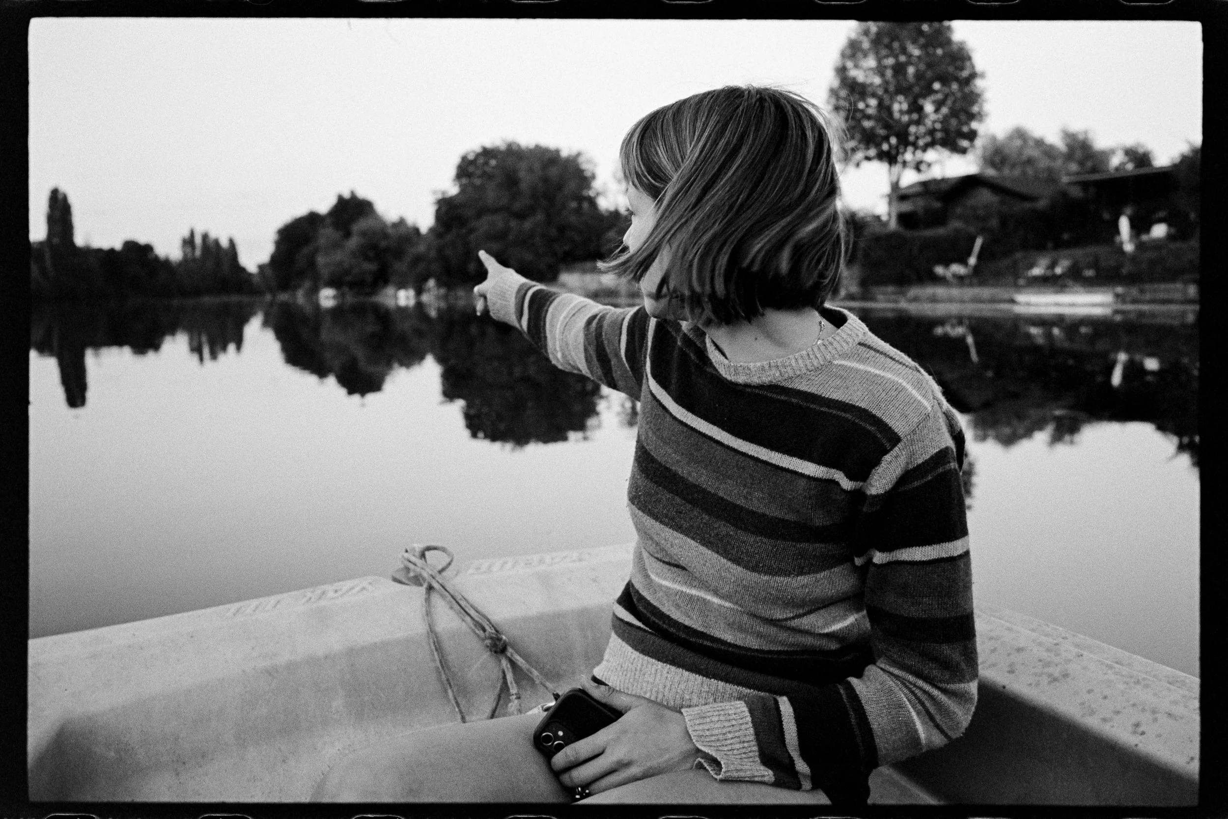 Une femme assise sur un bateau, pointant du doigt vers le lac devant elle, en arrière-plan arbres et maisons, en noir et blanc.