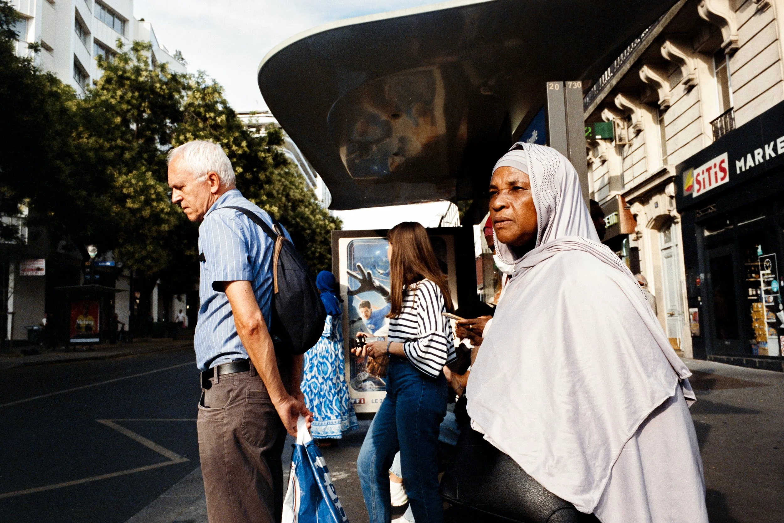 Gens attendant à un arrêt de bus dans une rue urbaine, avec des bâtiments et un panneau publicitaire en arrière-plan.