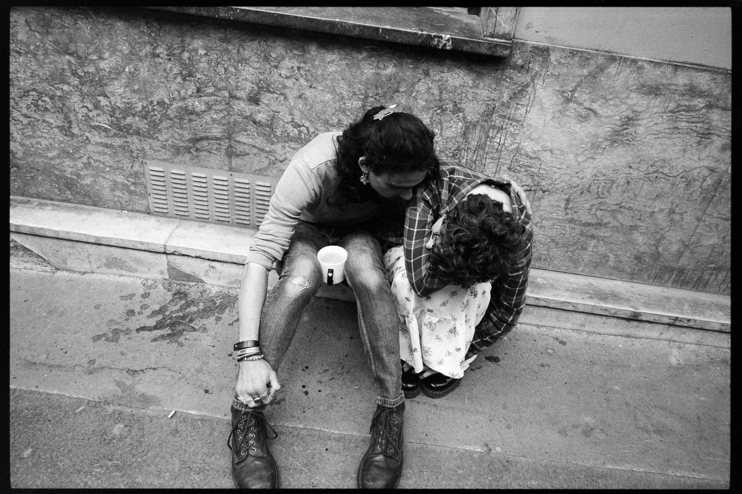 Deux femmes assises sur le trottoir, l'une aide l'autre à se relever, dans un décor urbain en noir et blanc.
