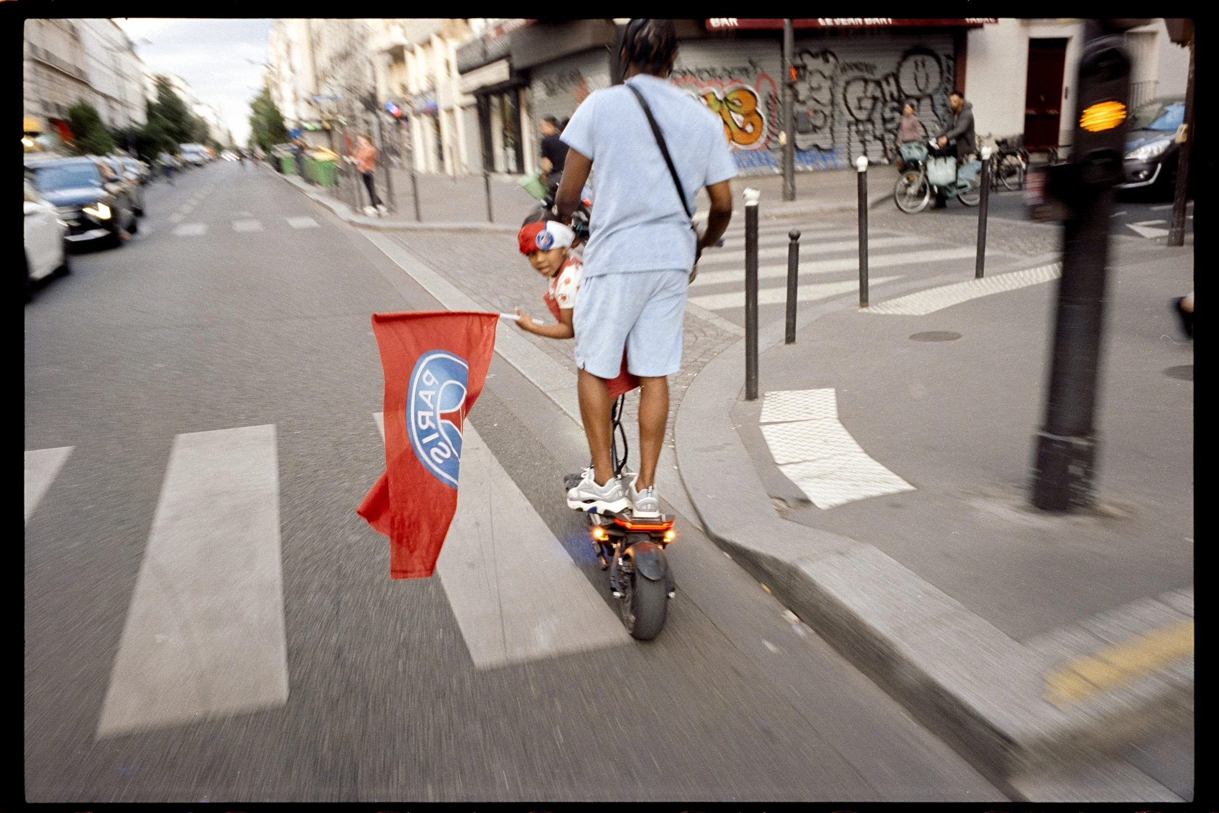 Une personne sur un hoverboard portant un enfant en hiérarchie, dans une rue urbaine avec des passants, des voitures et des graffitis sur le mur.