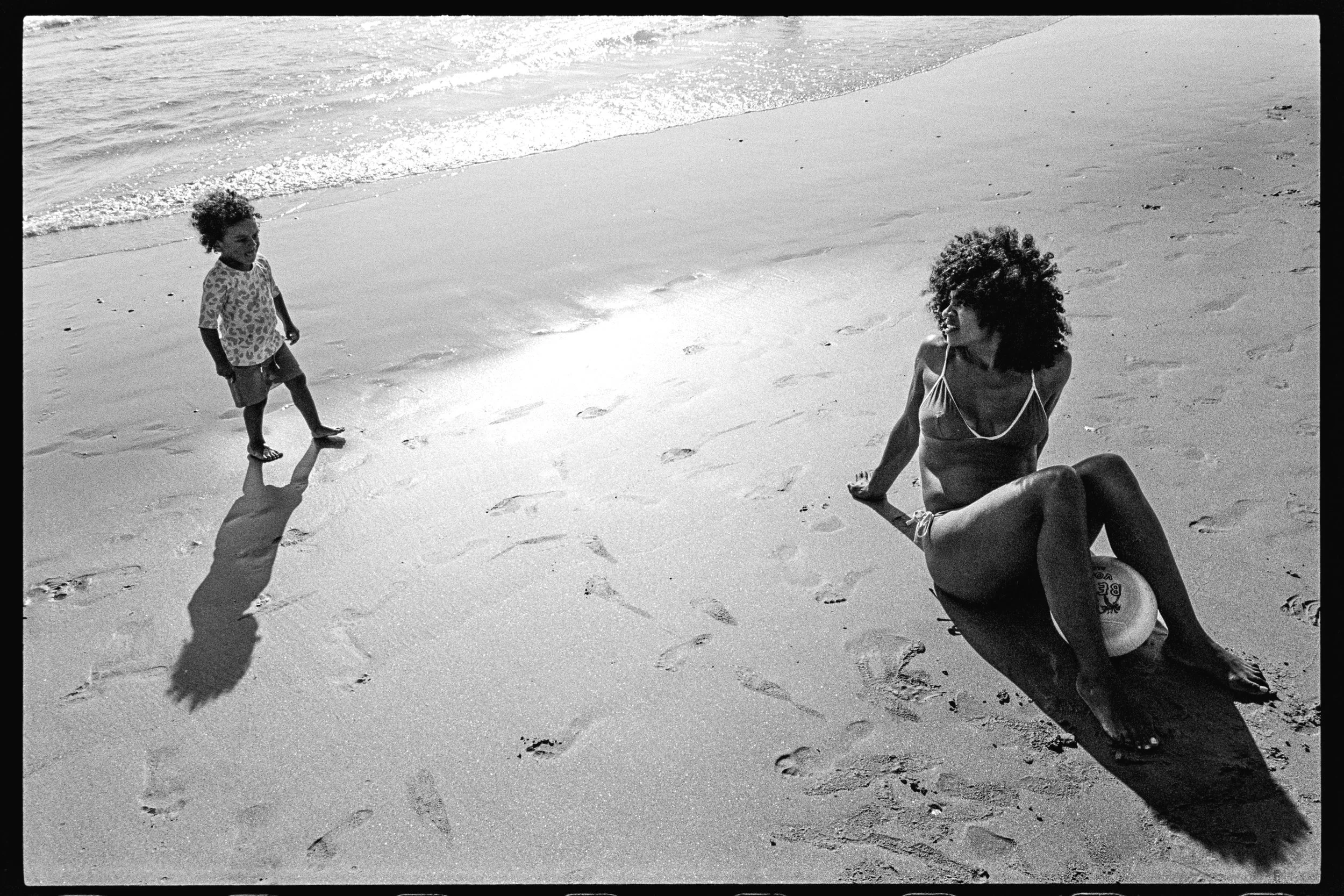 Une femme assise sur le sable d'une plage avec un ballon, regardant un enfant qui marche vers elle, dans une scène en noir et blanc.