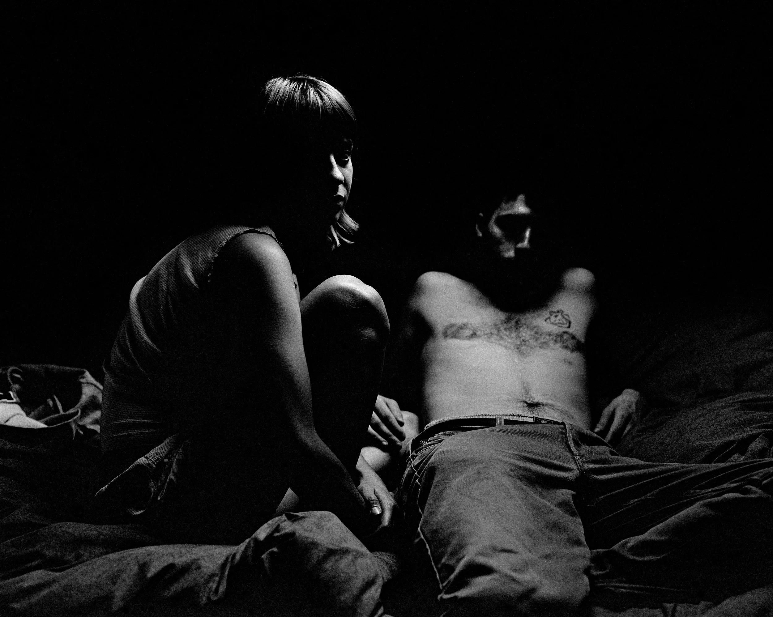 Une photo en noir et blanc de deux personnes, une femme assise en tailleur et un homme allongé, dans un espace sombre. La lumière met en évidence leurs silhouettes, mais leurs visages sont partiellement cachés dans l'ombre.