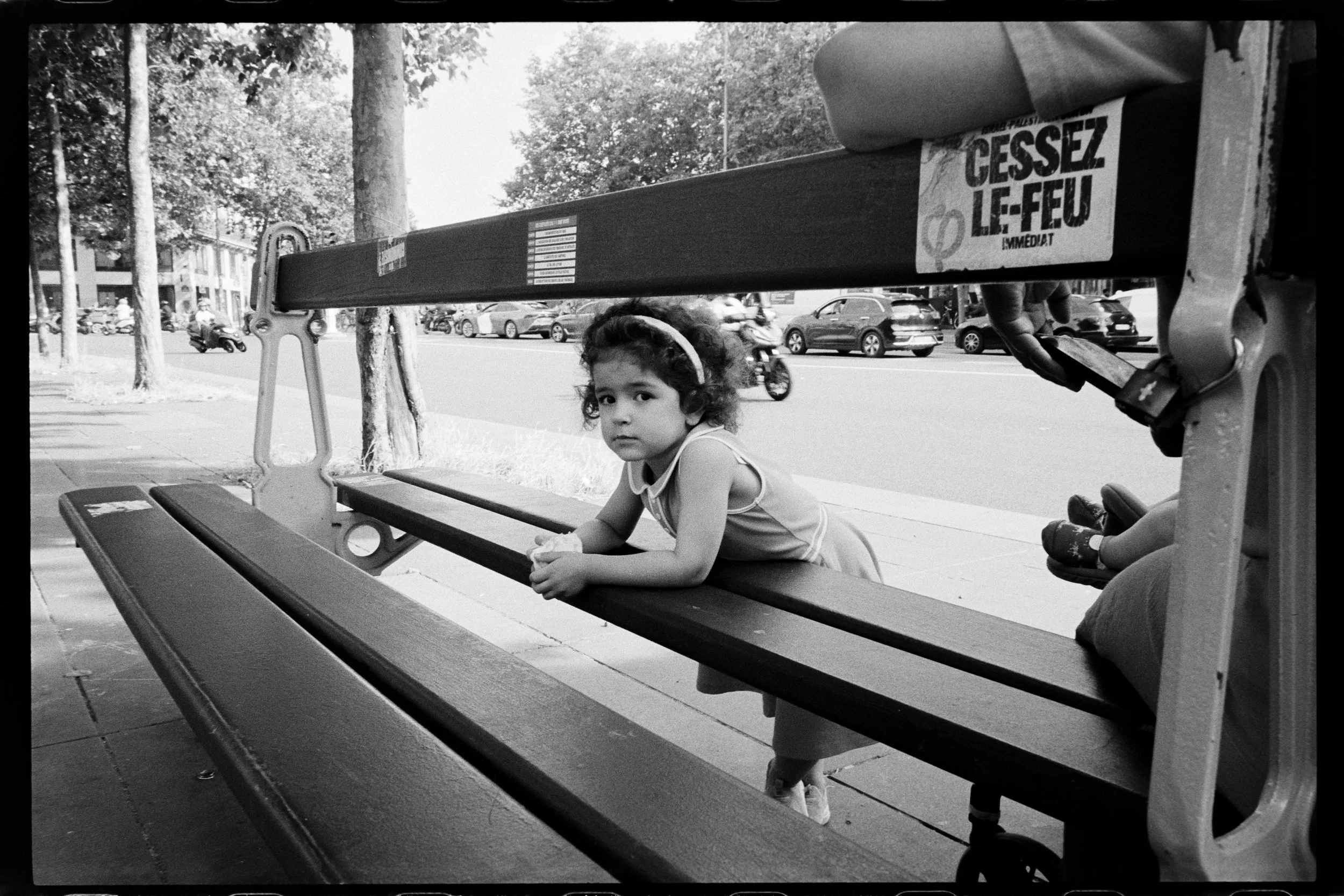 Une jeune fille blonde en robe est assise sur un banc public, regardant vers la caméra, dans une rue urbaine.