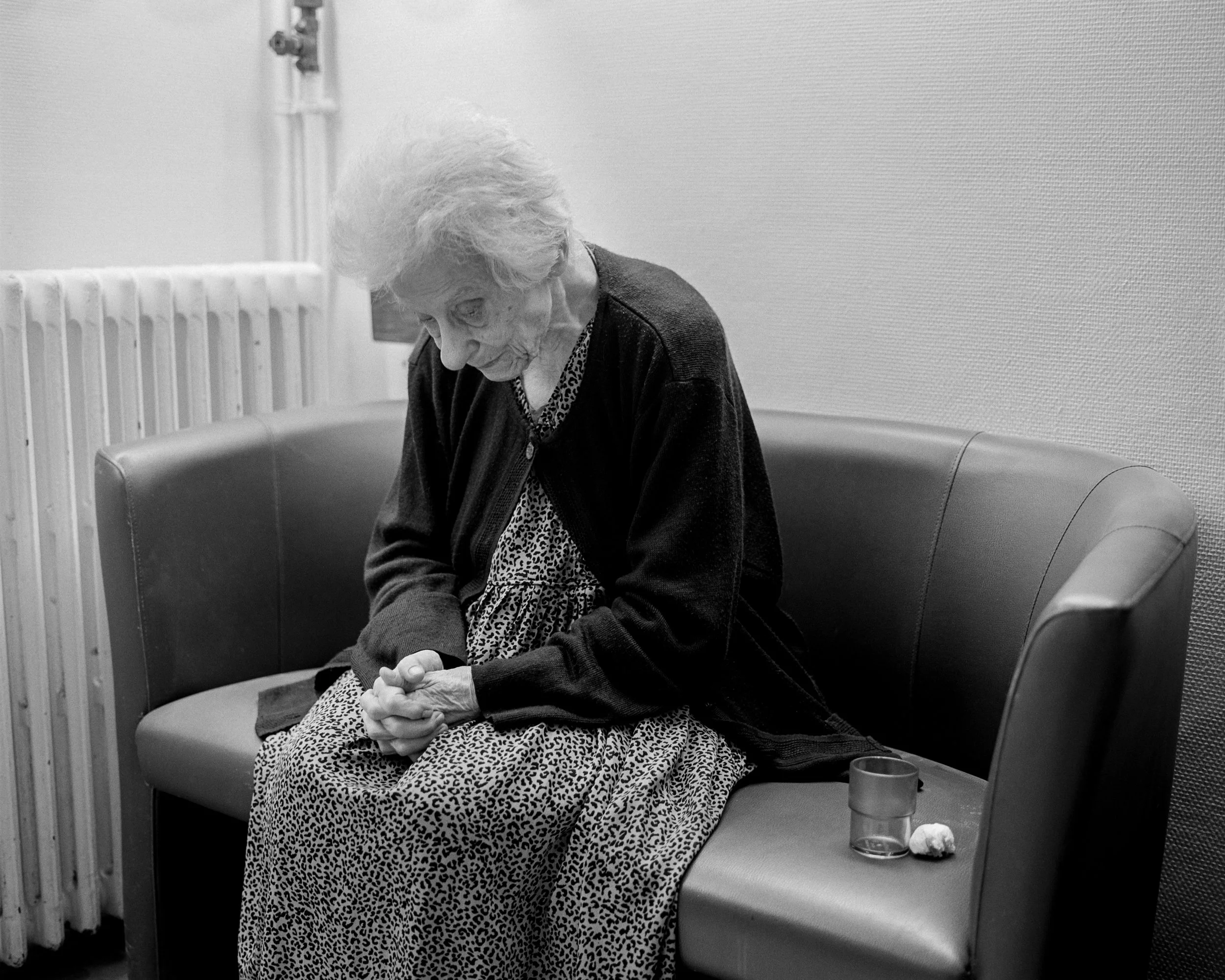 Une femme âgée assise sur un canapé, les mains jointes, dans une pièce aux murs clairs et un radiateur visible à gauche.