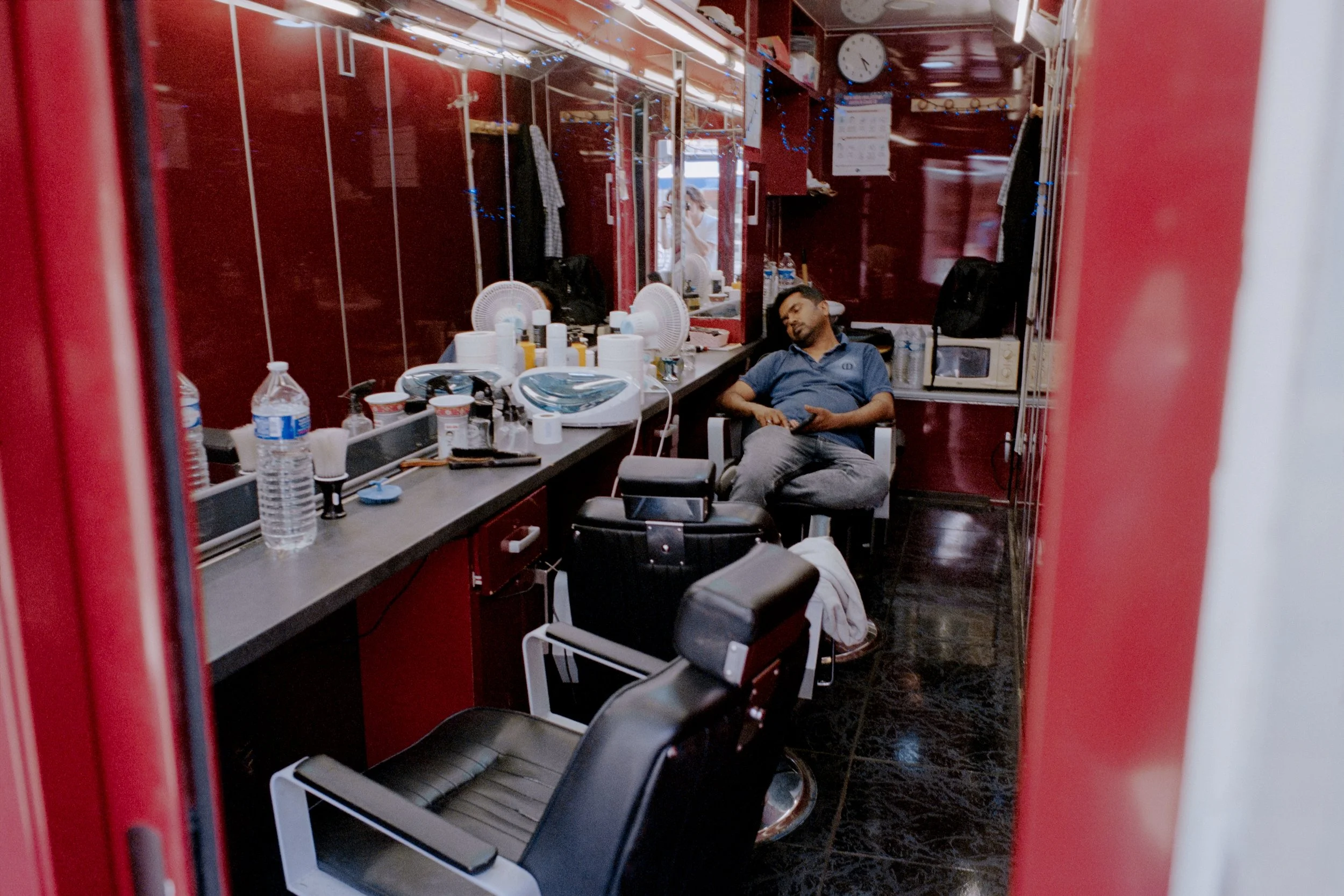 Un homme dormant dans un salon de barbier avec des fauteuils de barbier, des miroirs, et divers outils de coiffure sur le comptoir.
