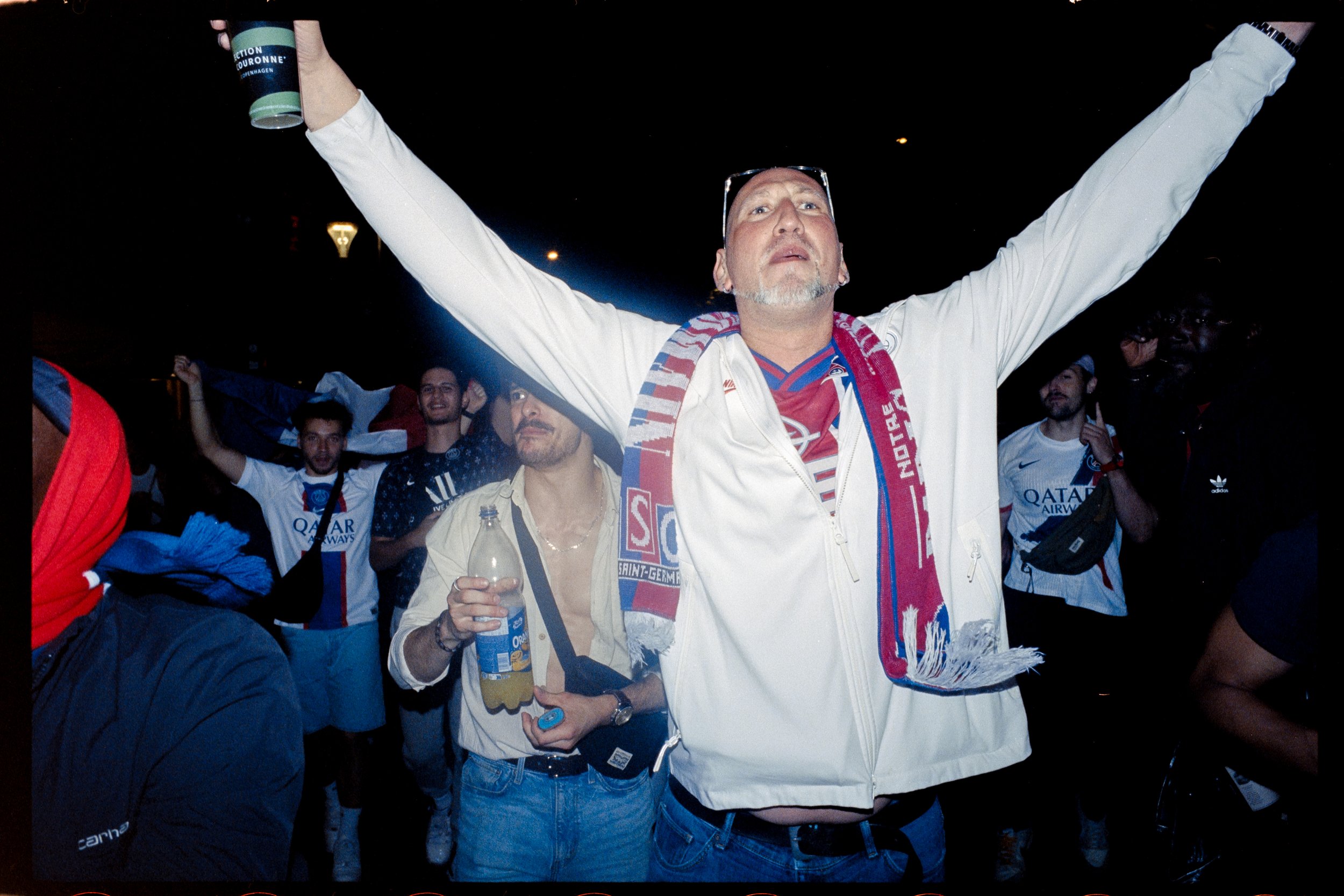Un groupe de supporters de football célébrant, portant des écharpes et des maillots de l'équipe, avec un homme au centre d arms levées, dans une ambiance festive.