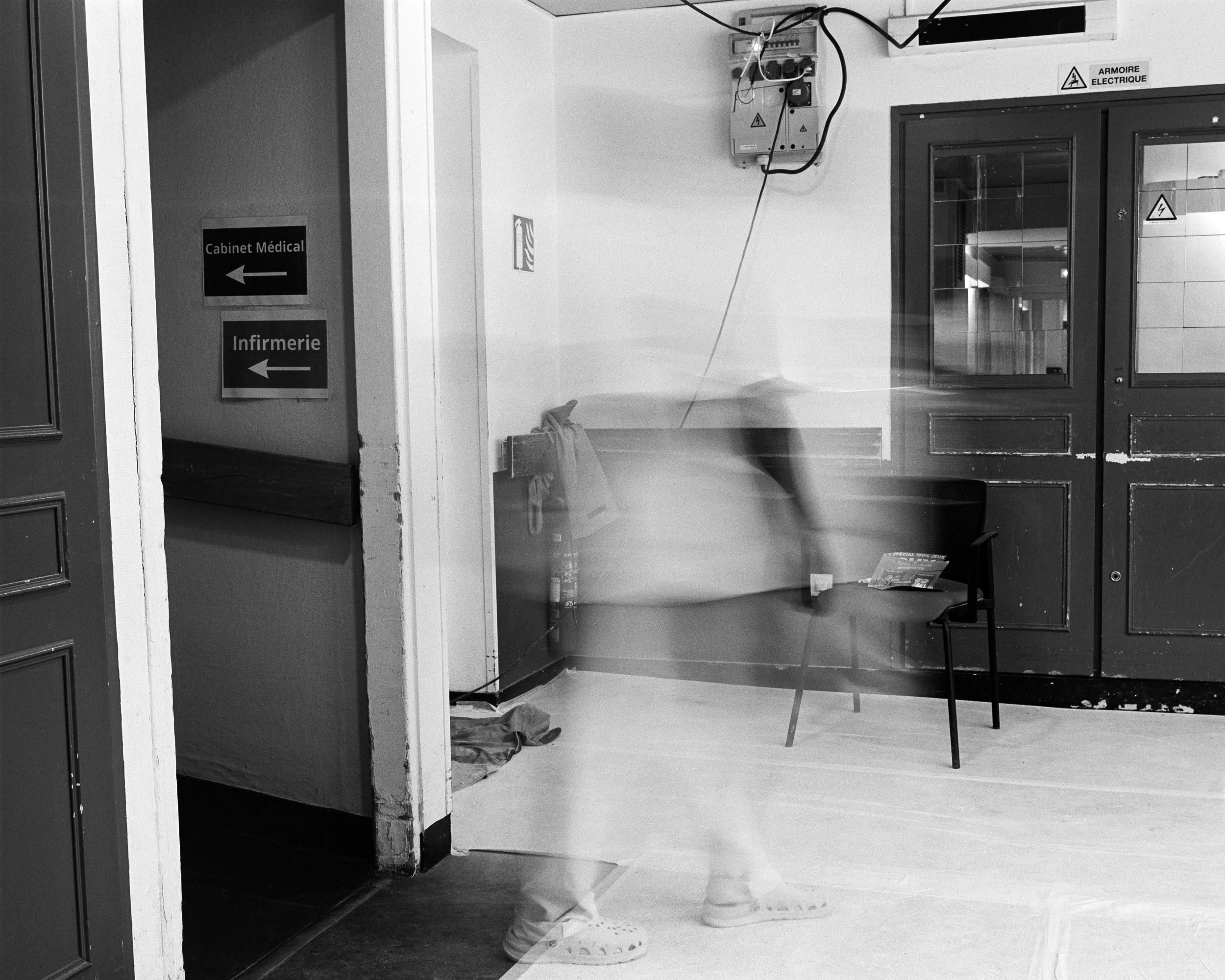 Intérieur d'un établissement médical avec une porte de salle d'attente, une chaise, et un panneau indiquant 'Cabinet Médical' et 'Infirmierie' avec flèches. Un patient ou visiteur passe rapidement devant la caméra, créant une image floue, en raison d