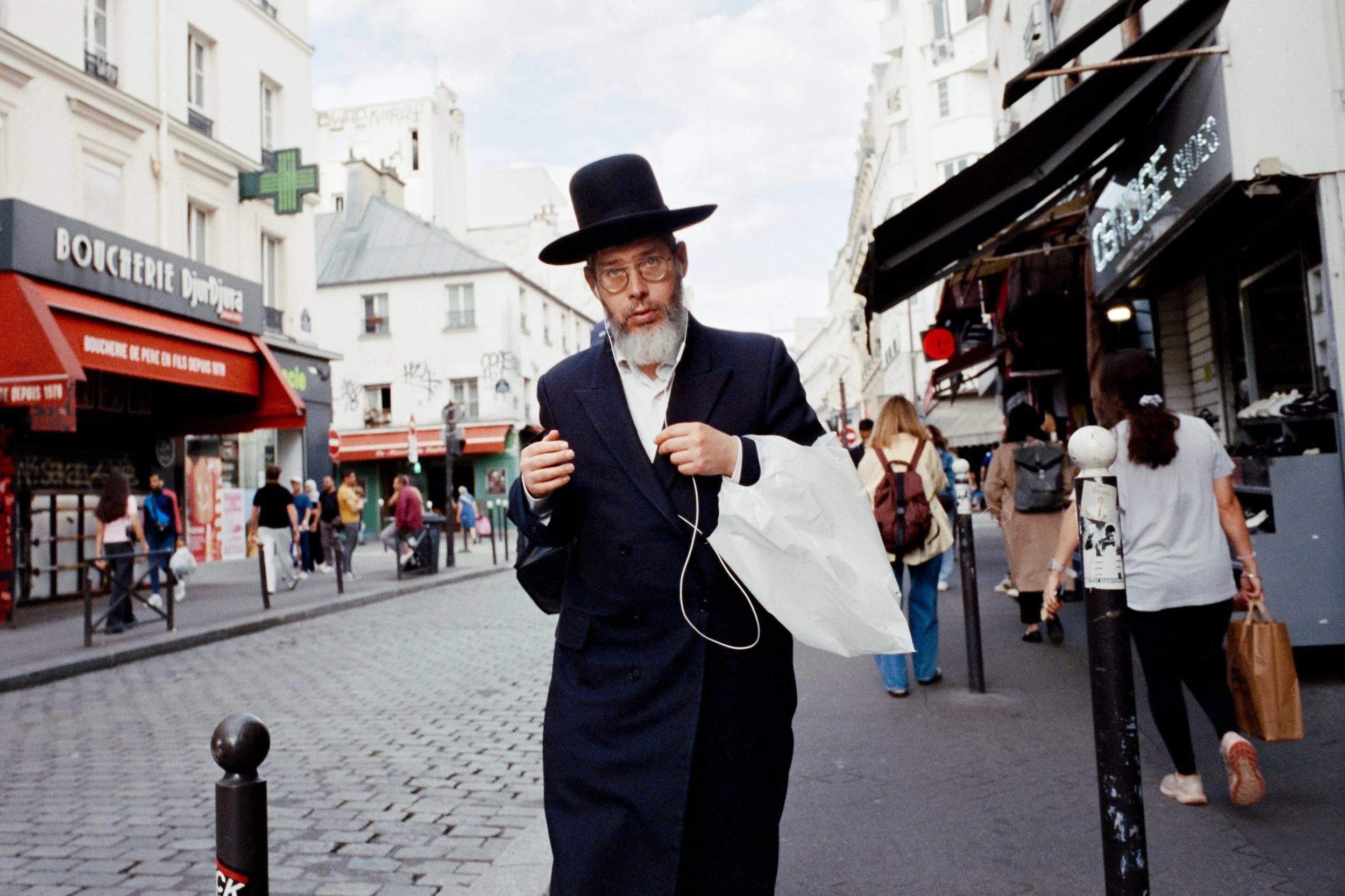 Un homme portant un costume noir, une chemise blanche, un chapeau noir, et des lunettes, marche dans une rue fréquentée de Paris, avec des magasins et des passants en arrière-plan.