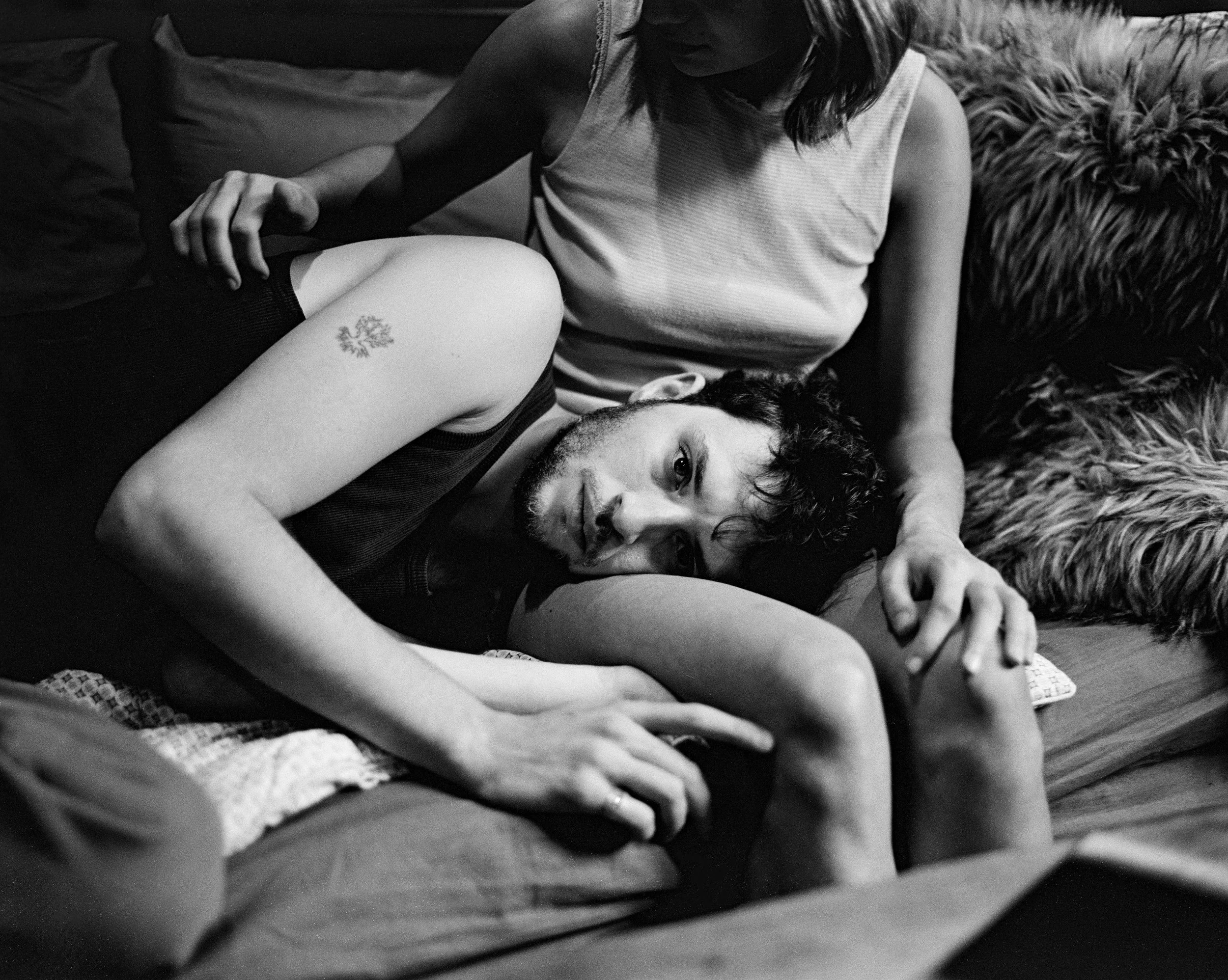 Un homme allongé sur un lit, regardant fixement l'objectif, une femme assise derrière lui, posant sa main sur son bras, dans une atmosphère intime.
