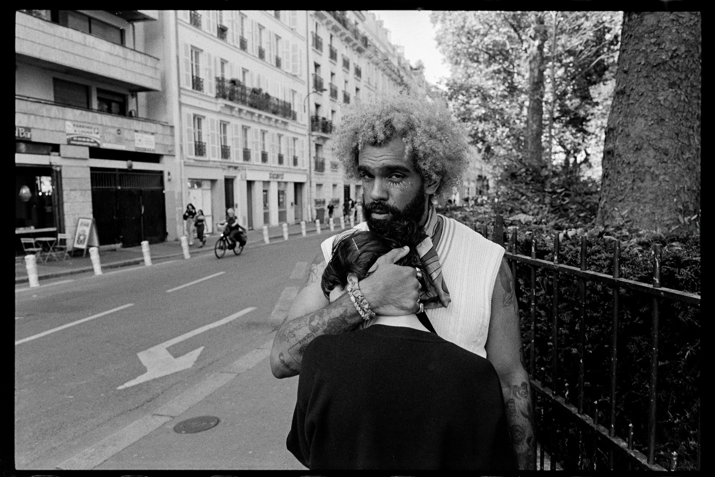 Deux personnes en interaction sur un trottoir d'une rue urbaine, l'une avec des cheveux bouclés frisottés et la peau foncée, l'autre avec des cheveux courts, assise, face à lui, entourée de bâtiments résidentiels.