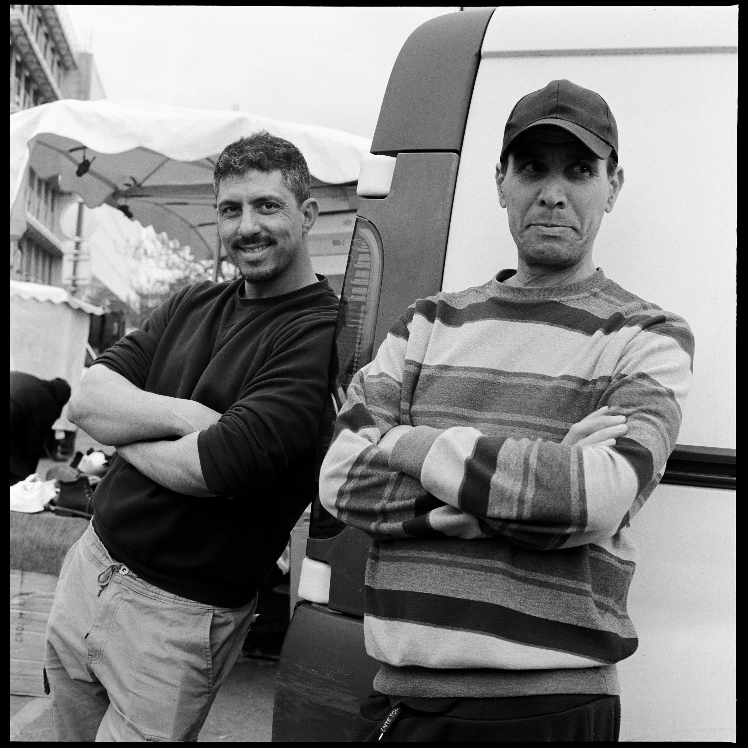 Deux hommes posent devant un camion dans une rue urbaine, l'un souriant et l'autre avec une expression sérieuse, tous deux ont les bras croisés.