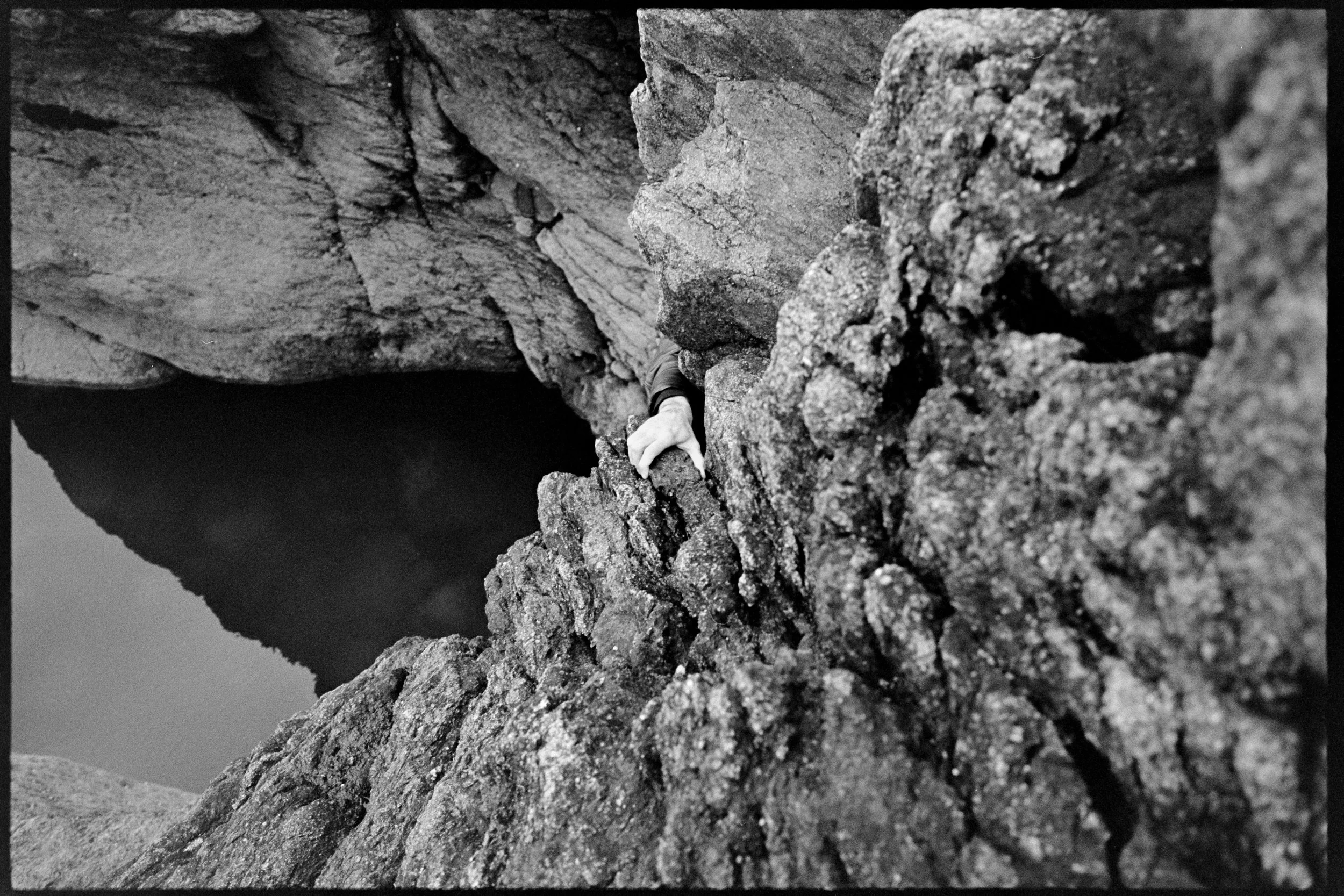 Une main attrape une roche sur une falaise rocheuse en bord de mer, avec une mer calme en arrière-plan.