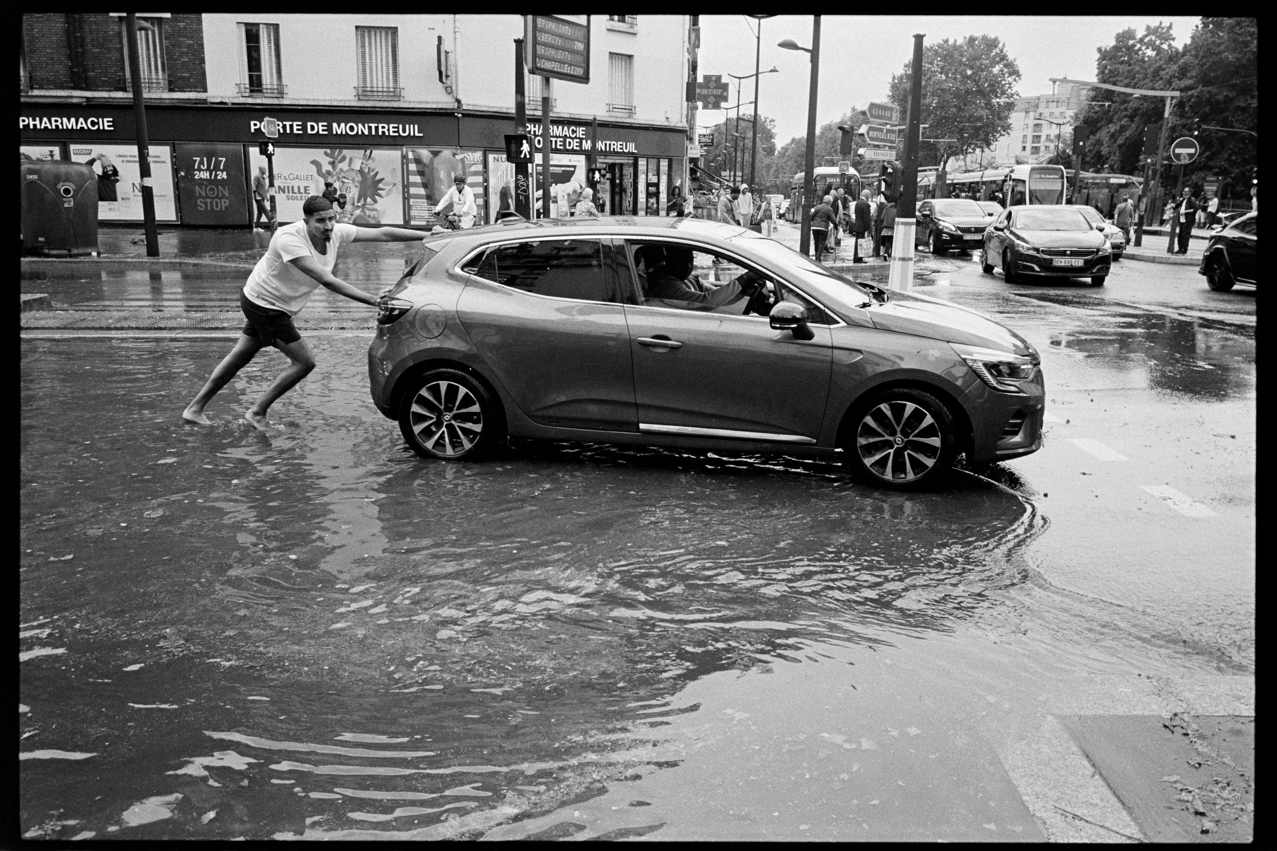 Une femme pousse une voiture garée sous la pluie dans une rue urbaine, avec des passants et des véhicules autour.
