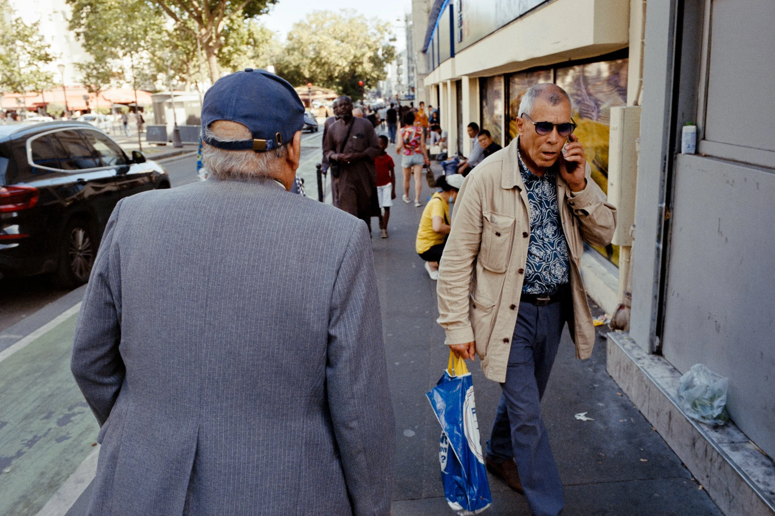 Deux hommes âgés, dont un porte une veste en tweed et un chapeau bleu, discutent dans une rue animée. L'autre porte une veste beige, des lunettes de soleil, et parle au téléphone tout en portant un sac en plastique bleu