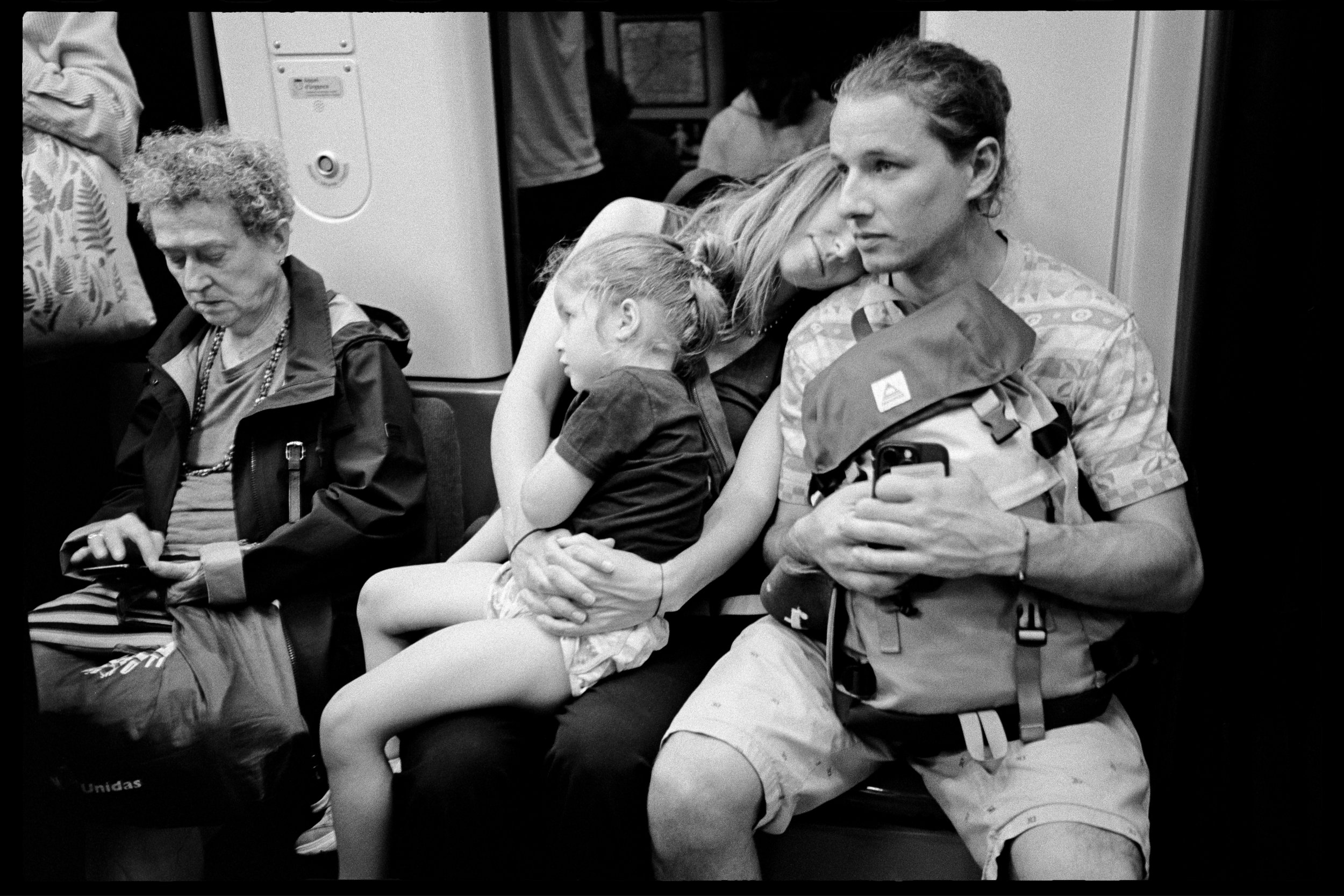 Quatre personnes assises dans un train, dont un homme avec un sac à dos, une femme avec une enfant endormie, et une femme âgée regardant son téléphone.