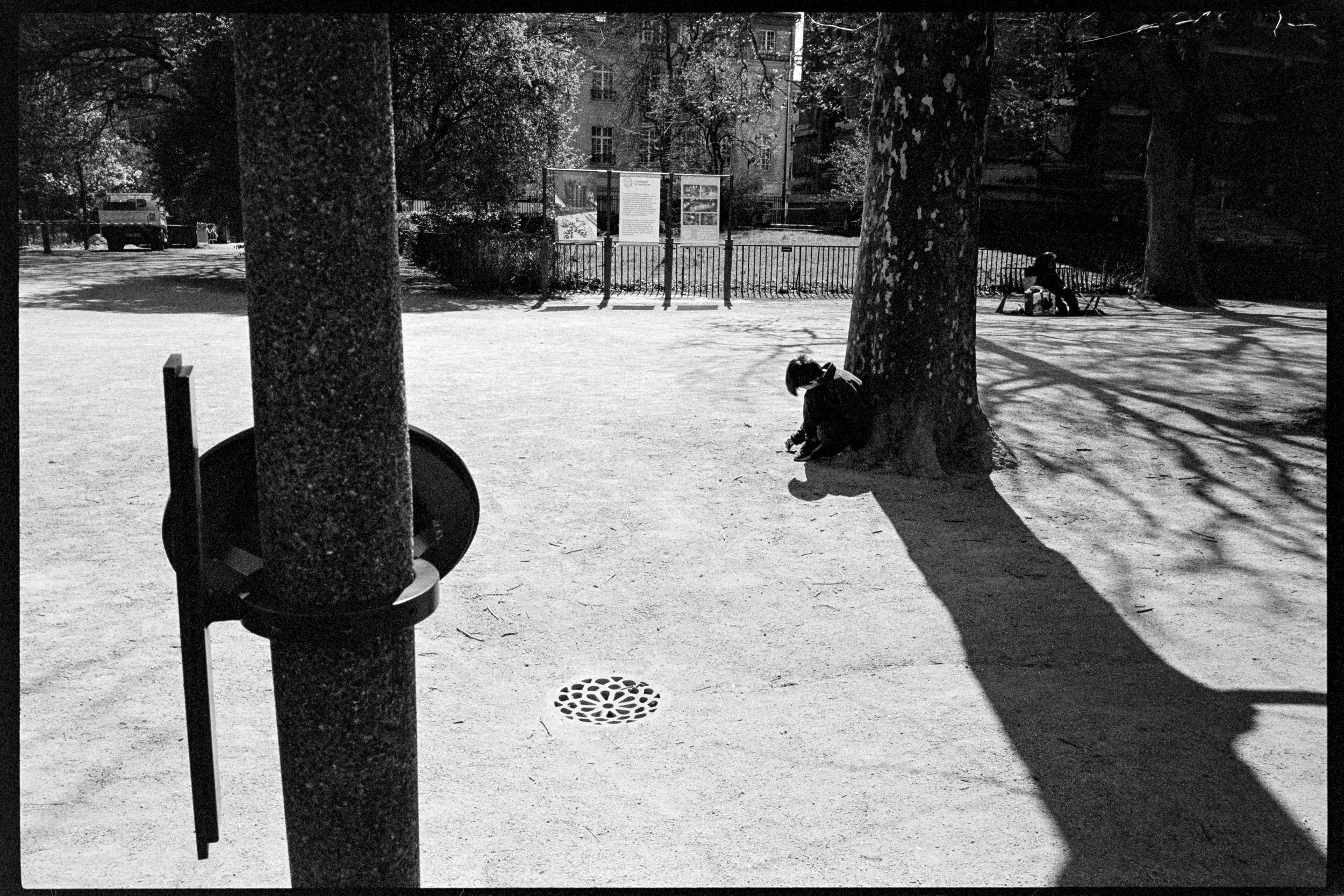 Une scène dans un parc avec deux personnes assises sous des arbres et des ombres longues, en noir et blanc.
