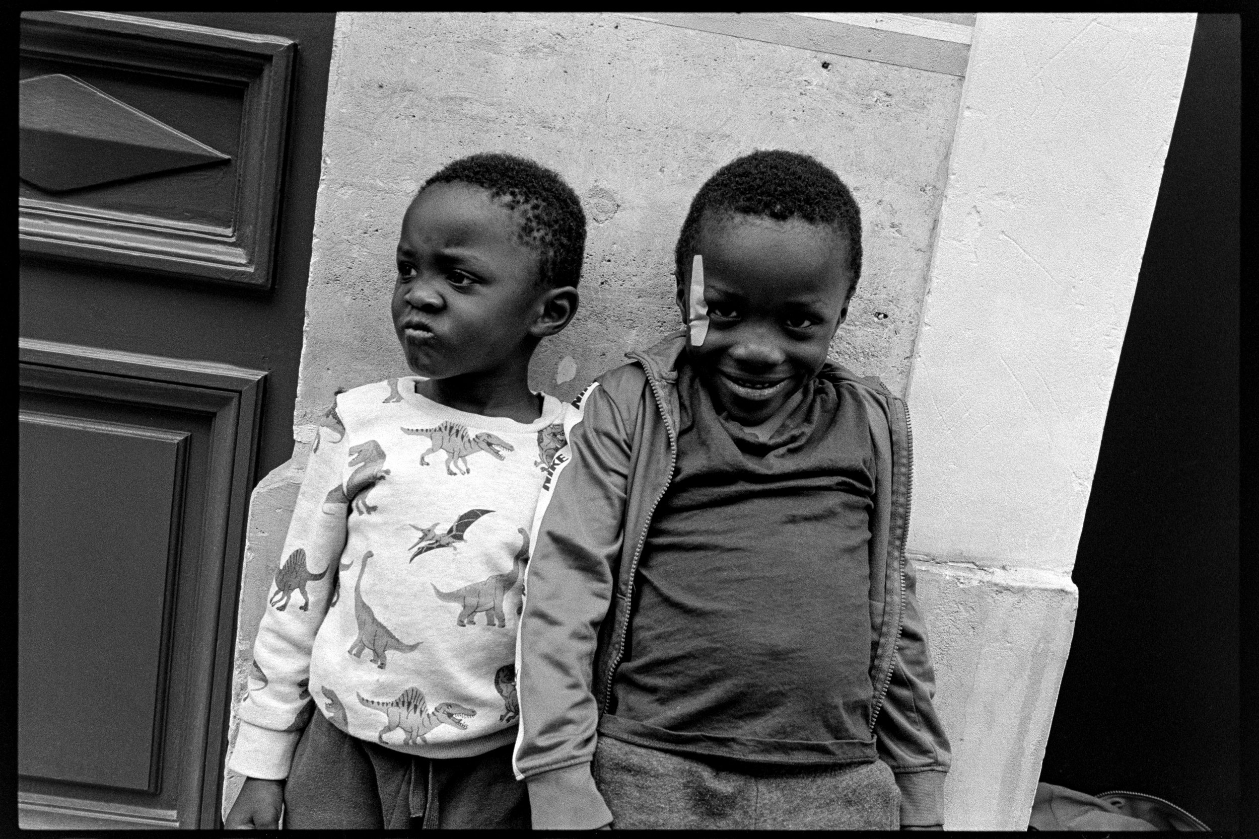Deux jeunes garçons noirs posant contre un mur, l'un porte un sweat à motifs dinosaures, l'autre un t-shirt sombre avec une veste. L'un d'eux a une bande de tissu blanc sur le visage. La photo est en noir et blanc.