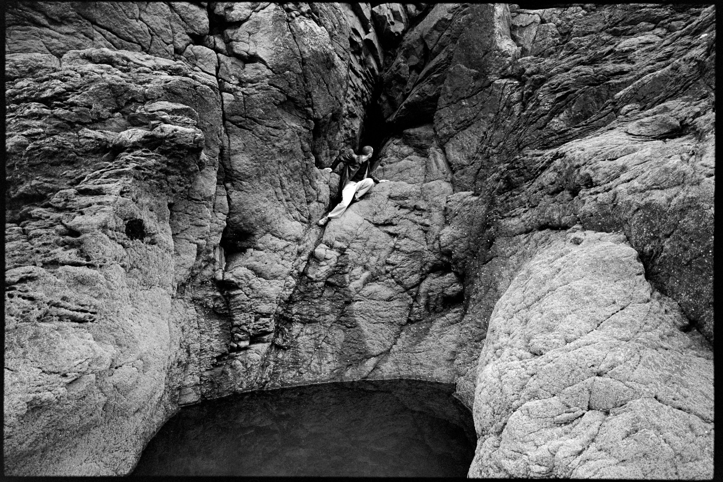 Une personne escalade une paroi rocheuse dans une nature sauvage, avec un petit plan d'eau en bas et des rochers environnants.