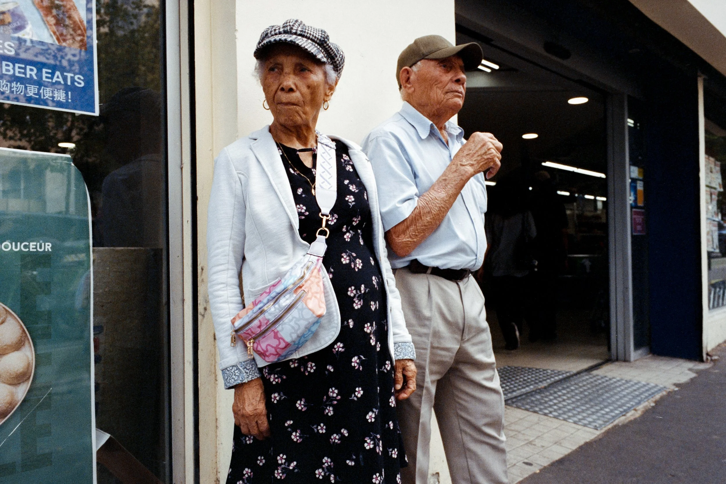 Une femme âgée et un homme âgé se tiennent devant un magasin, tous deux portent des casquettes et ont une expression sérieuse.