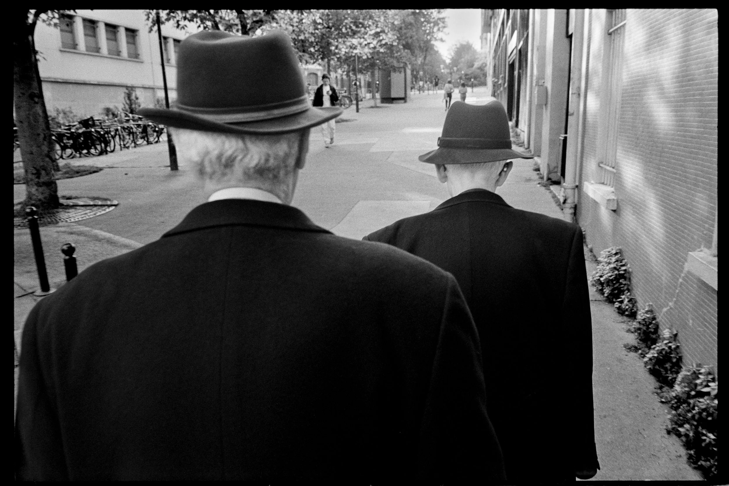 Deux hommes en costume noir et chapeaux traversant une rue en ville, photographiés de dos, en noir et blanc.