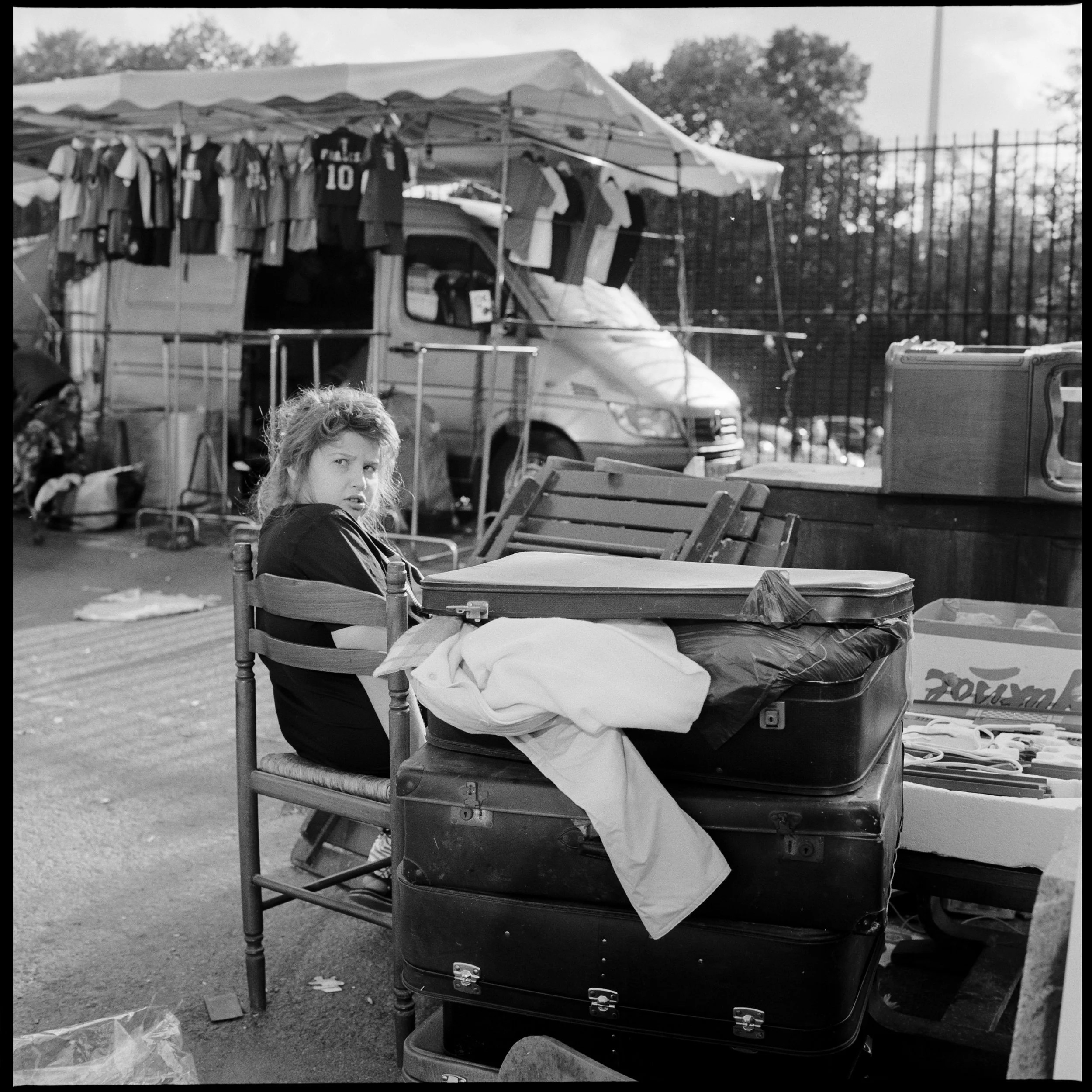 Une jeune femme assise à une table en plein air, avec des valises empilées et un stand de vêtements en arrière-plan.