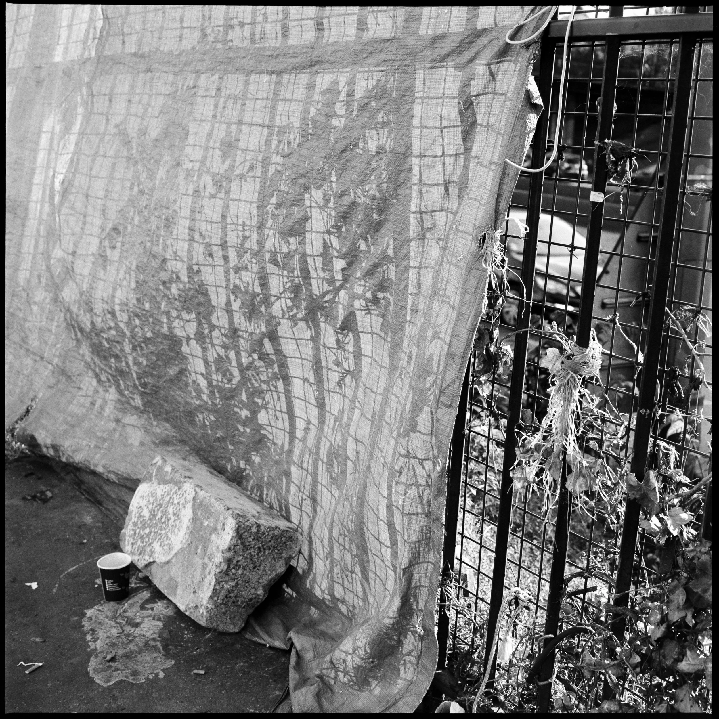 Une toile transparente, probablement une vieille toile de jute ou un tissu usé, tendue contre une clôture métallique. Sur le sol, il y a une brique posée sur un sac en plastique et un gobelet en plastique noir à côté.