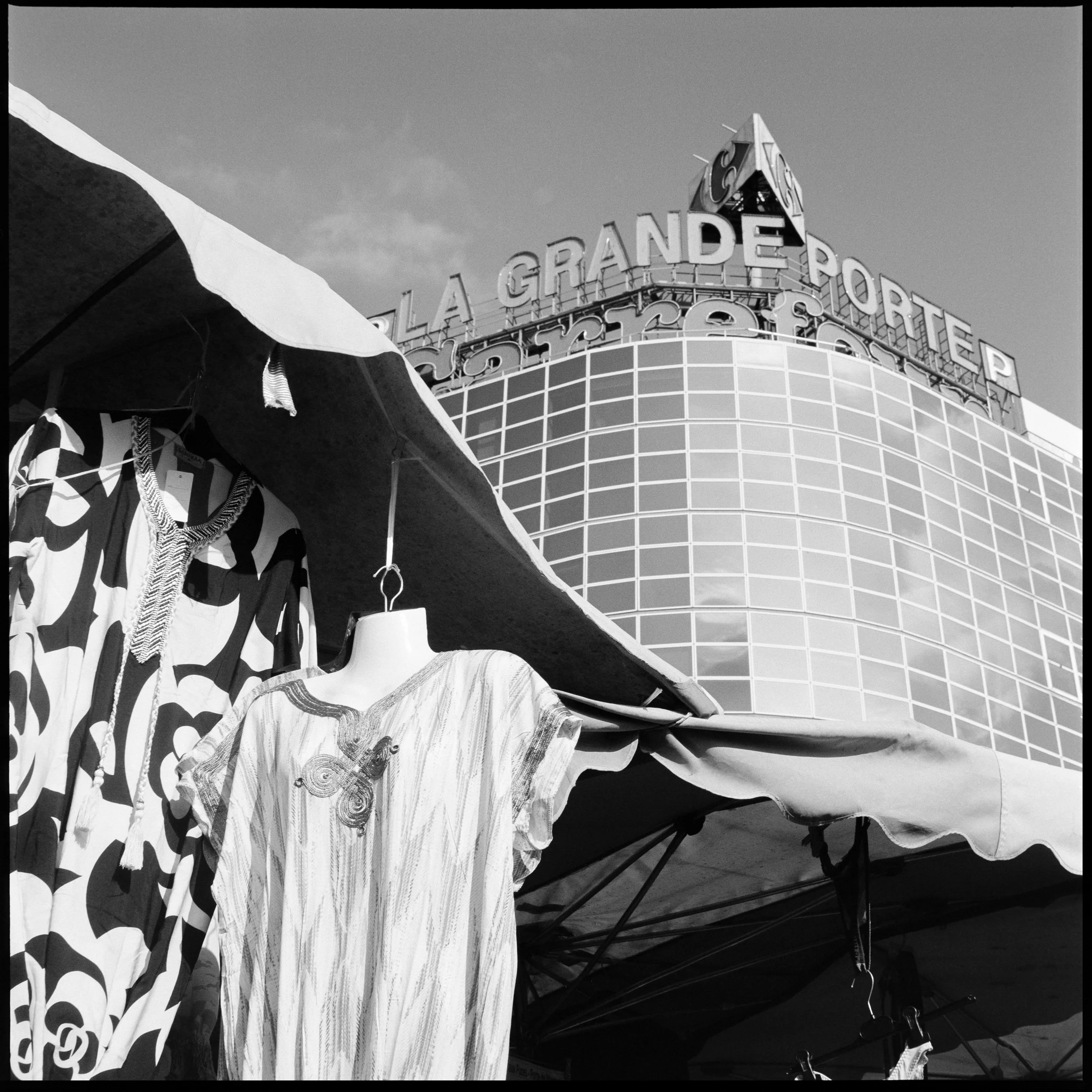 Vente de vêtements sous un stand de marché devant un bâtiment avec une enseigne en lettres capitales "LA GRANDE PORTER".