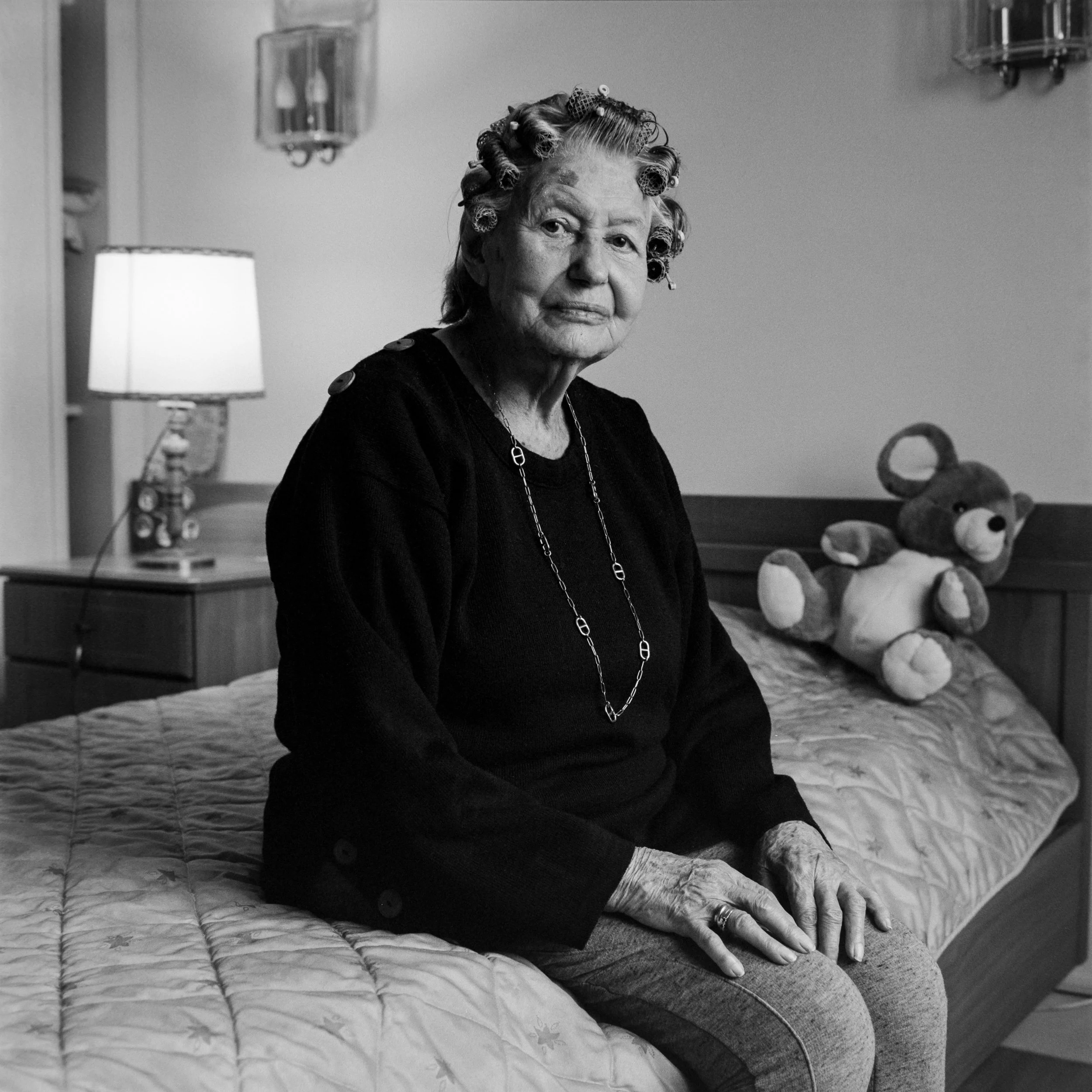 Une femme âgée avec des cheveux enroulés dans des rouleaux, assise sur un lit dans une chambre, avec une lampe de chevet allumée et un ours en peluche sur le lit.