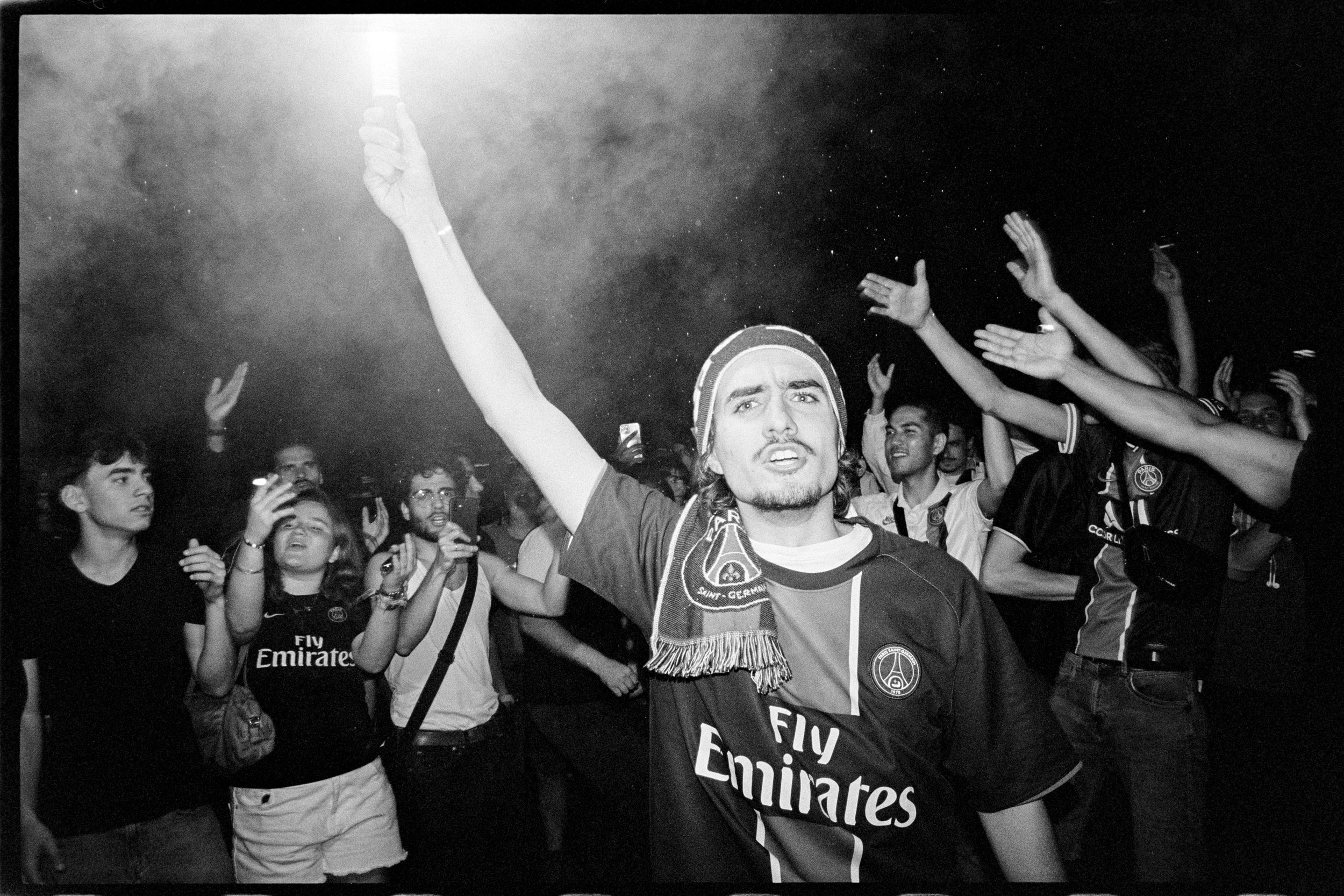 Un groupe de fans de football, dont un homme portant un maillot du Paris Saint-Germain et une écharpe, célèbre lors d'une manifestation nocturne, avec des bras levés et des expressions de joie et d'excitation.