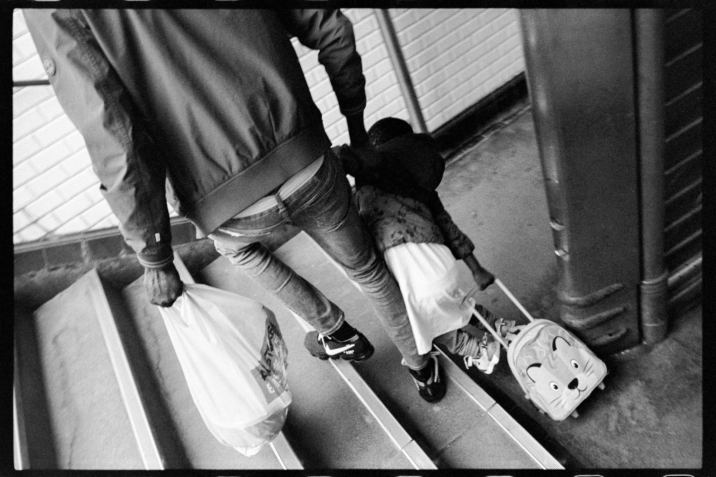 Une personne adulte portant un sac à main et un sac plastique, tenant la main d'un jeune enfant avec une valise à roulettes à l'effigie d'un visage de chat, dans un couloir avec des murs de briques et un sol en carrelage.