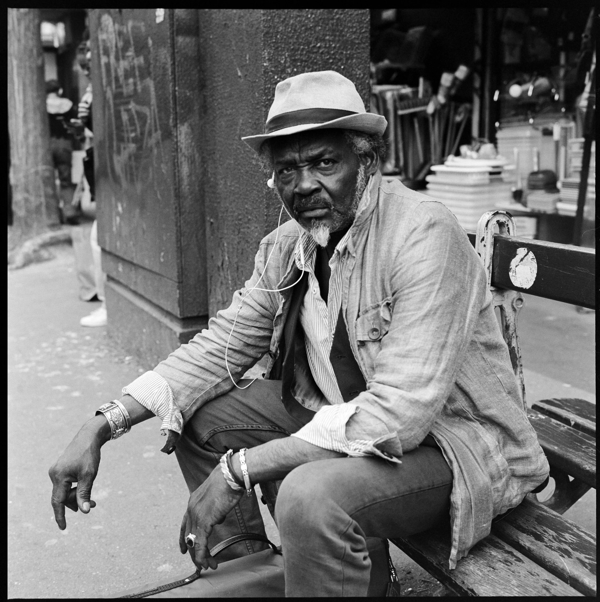 Un homme âgé assis sur un banc dans une rue urbaine, portant un chapeau et des vêtements décontractés, avec des écouteurs dans les oreilles, regardant fixement vers l'appareil photo. En arrière-plan, il y a un étal ou une boutique avec des livres ou 