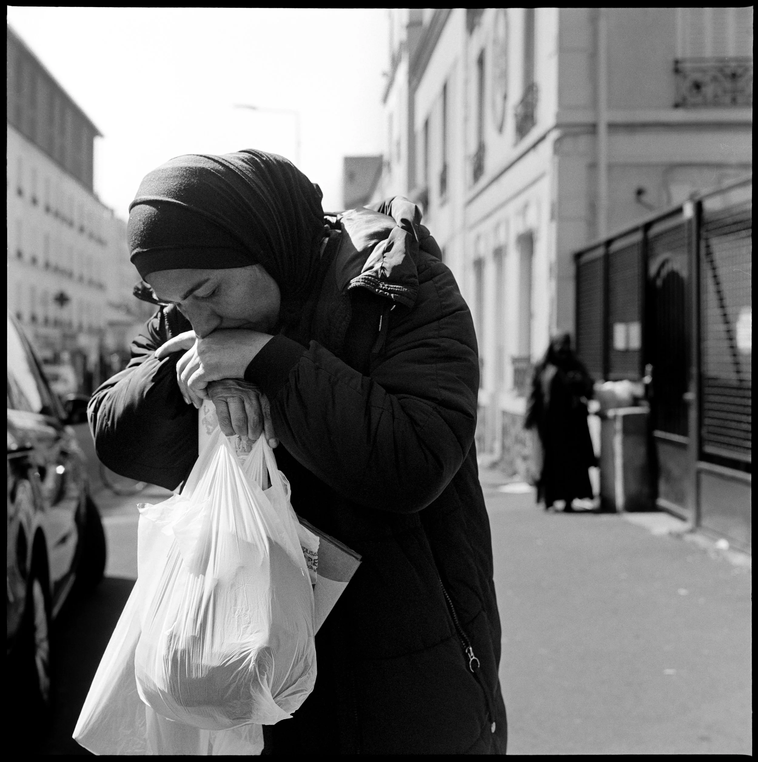 Une femme portant un hijab noire a les yeux baissés et semble en pleurs, tenant un sac en plastique avec ses mains jointes, dans une rue urbaine. En arrière-plan, d'autres personnes sont présentes près d'une grille ou d'une clôture.