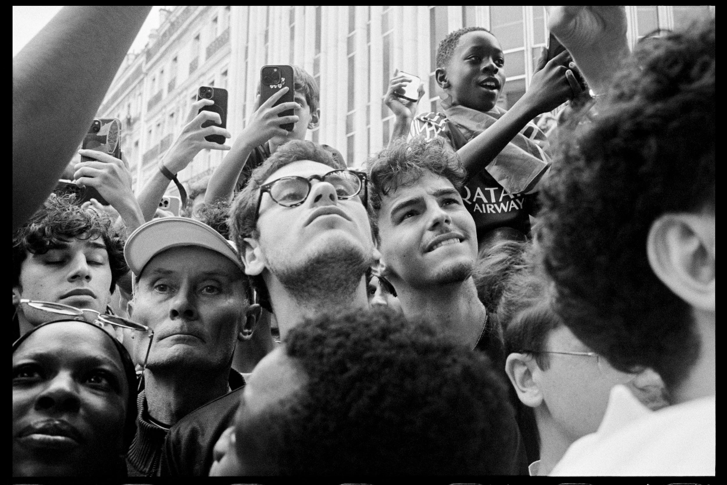 Gros groupe de personnes lors d'une manifestation ou rassemblement en milieu urbain, certains prenant des photos ou vidéos avec leurs téléphones, visage sérieux ou concentré.