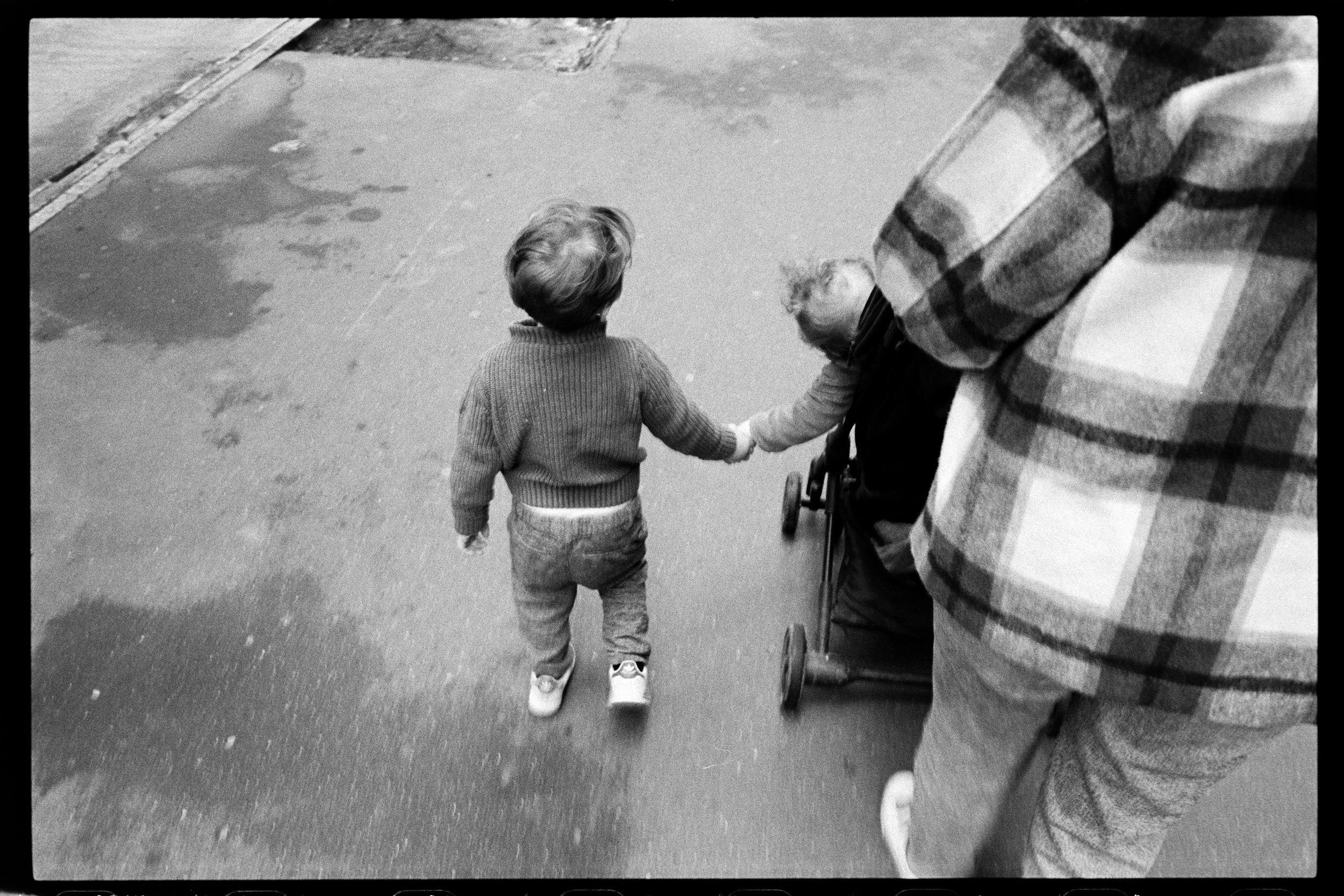 Un jeune garçon marche main dans la main avec une personne âgée en poussette, une autre personne marche à côté, sur un trottoir.