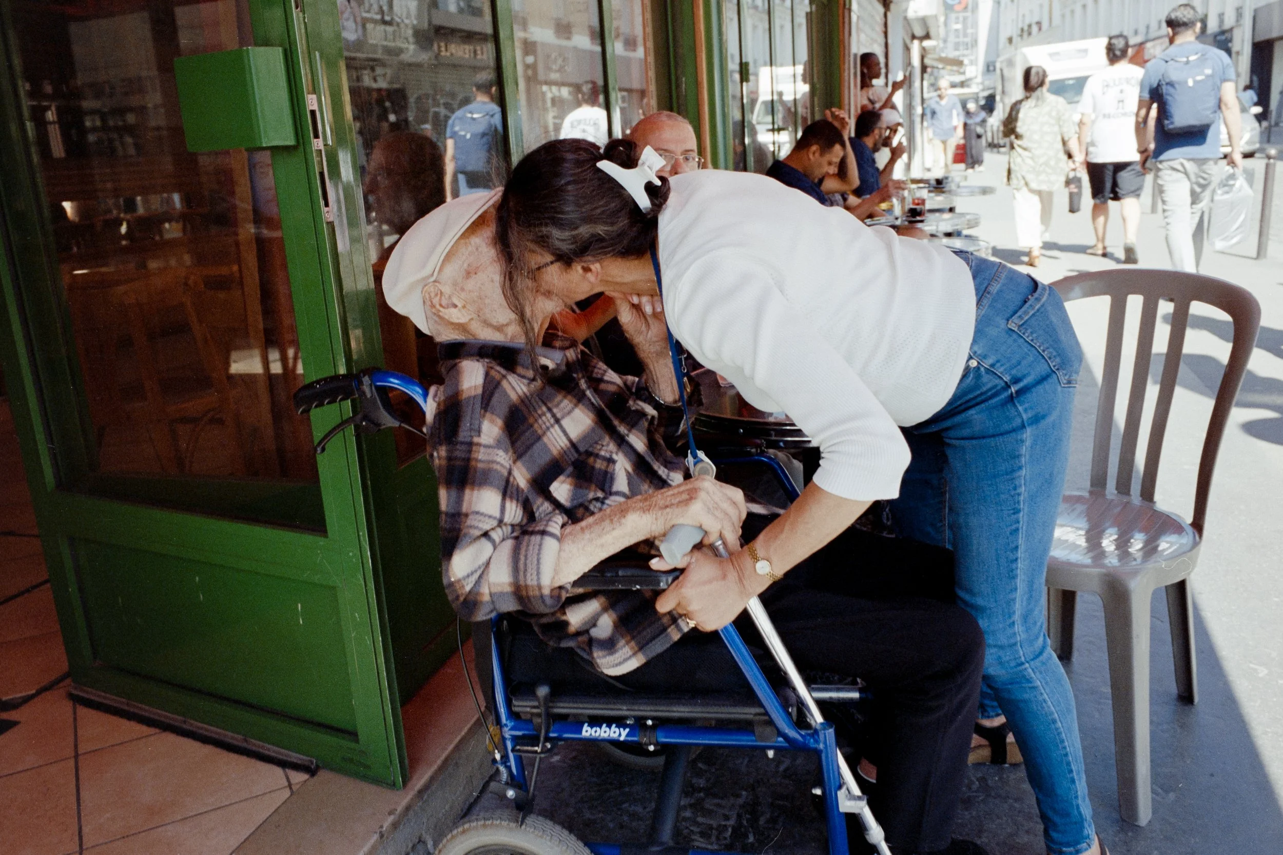 Une jeune femme aidant une personne âgée en fauteuil roulant à sortir d'un café en discutant, dans une rue animée.