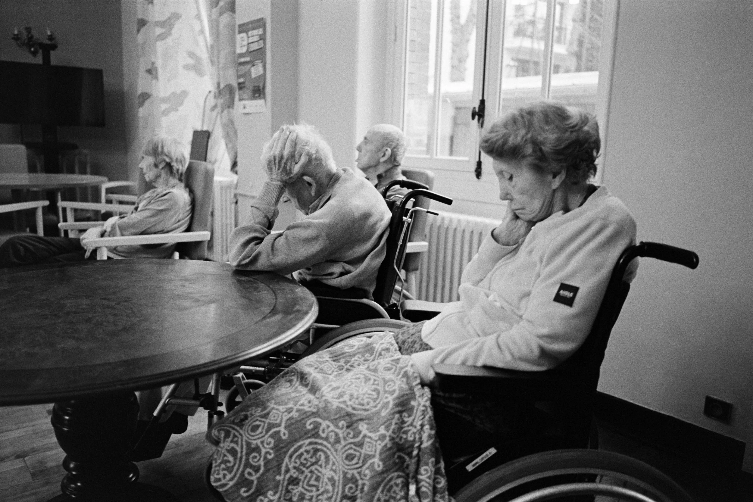 Quatre personnes âgées, dont trois en fauteuil roulant, assises dans une salle, semblant fatiguées ou pensives, avec une fenêtre en arrière-plan.