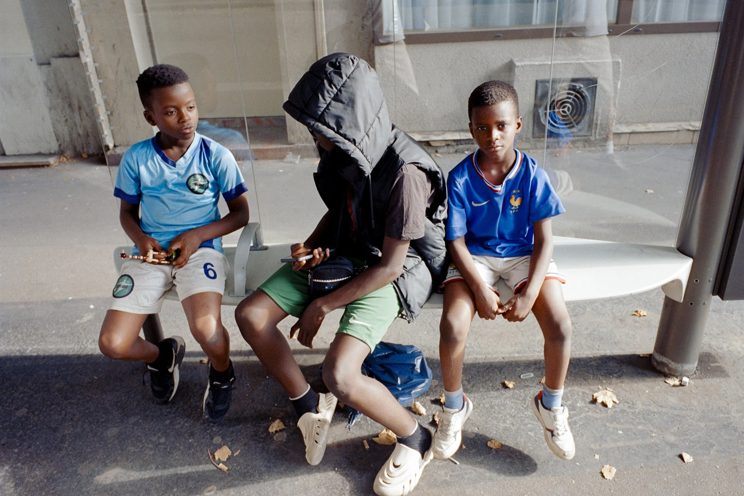 Trois enfants assis sur un banc à une station de bus, deux garçons portent des maillots de sport bleus et un garçon avec un sweat à capuche et un sac à bandoulière regardant son téléphone.