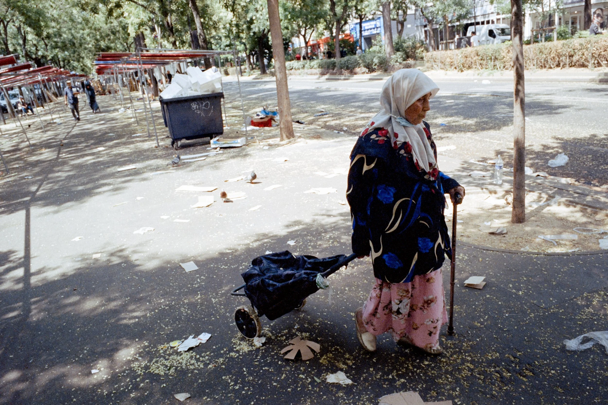 Une vieille femme marche dans une rue sale, tirant un chariot avec ses effets personnels, entourée de débris et de papiers.