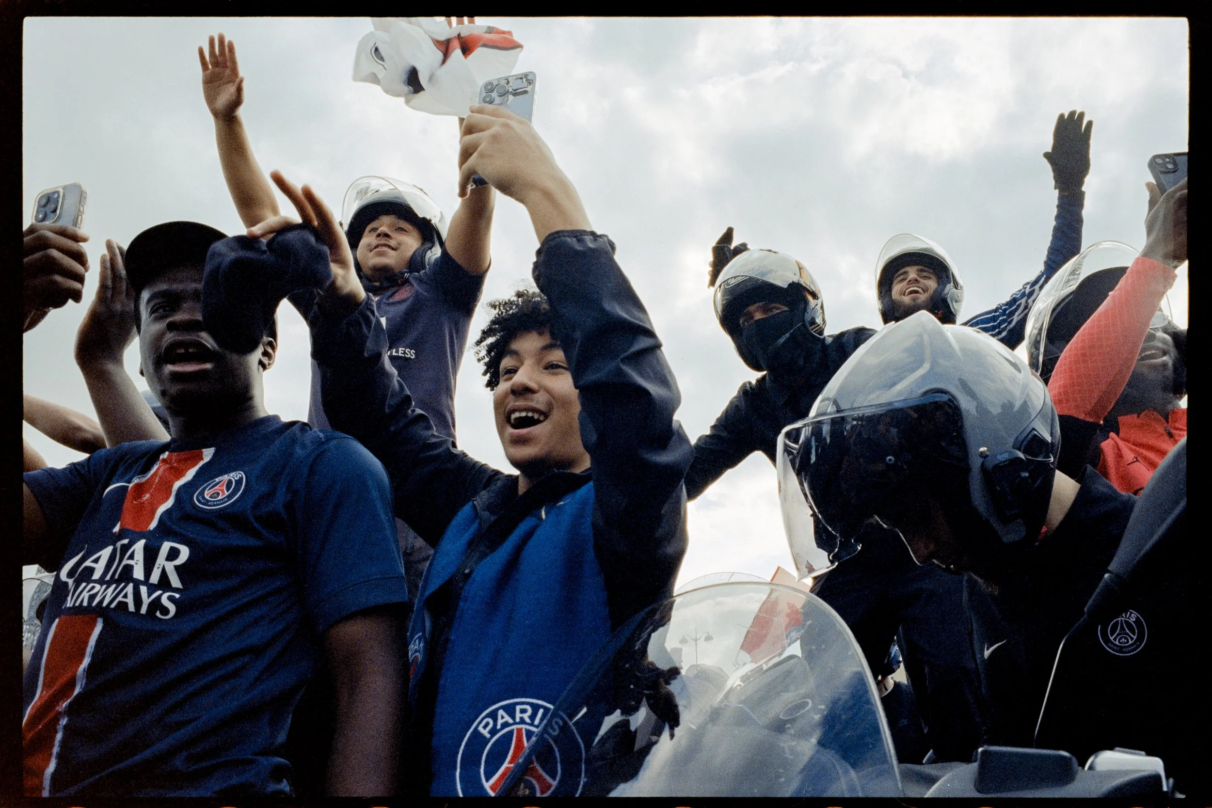 Groupe de jeunes, certains portant des casques de moto, heureux et en train de prendre des selfies, devant un ciel nuageux.