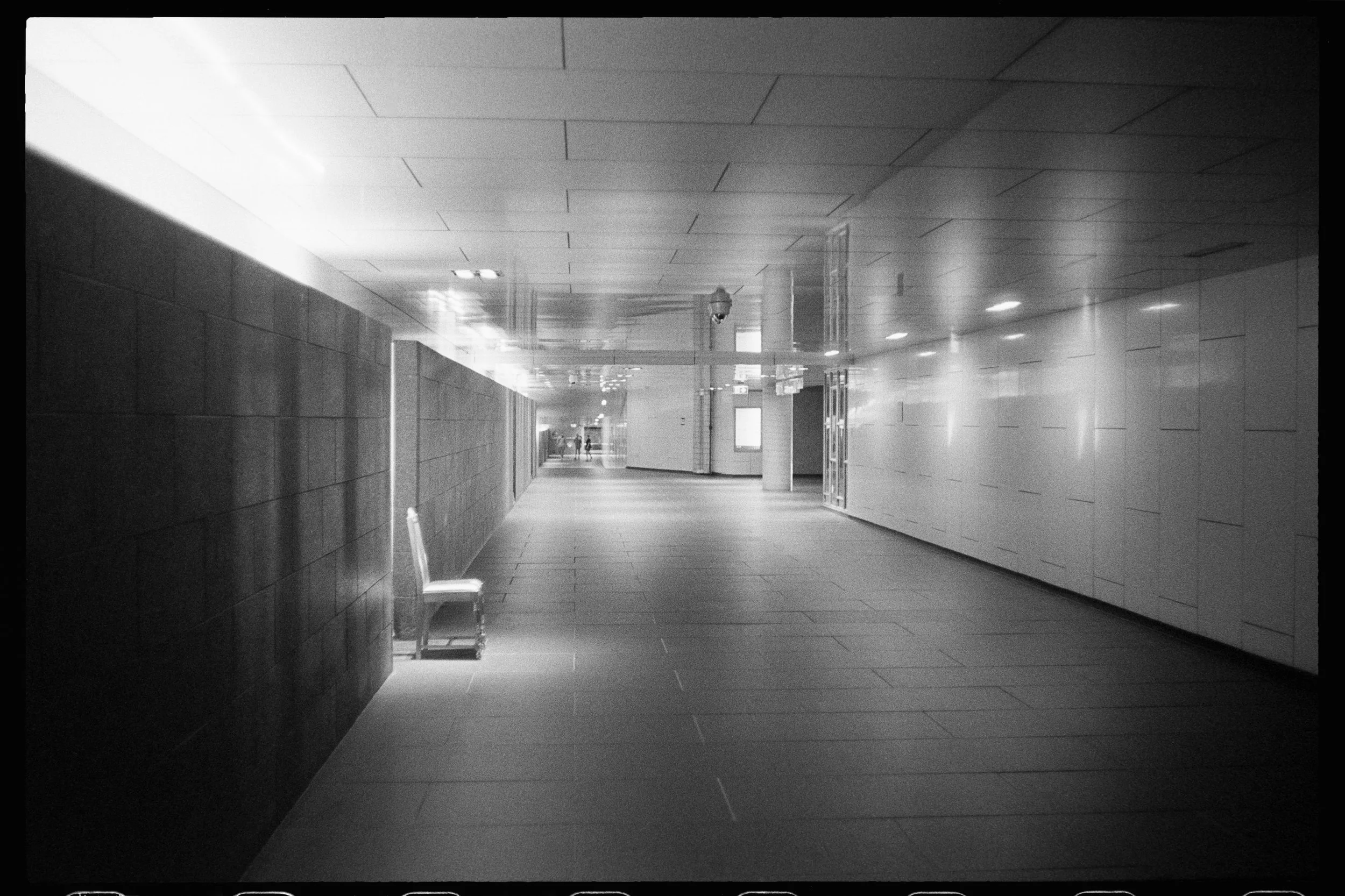 couloir d'un bâtiment moderne avec des murs en pierre, un sol en carrelage, des plafonds suspendus, une banquette blanche et plusieurs personnes au fond.
