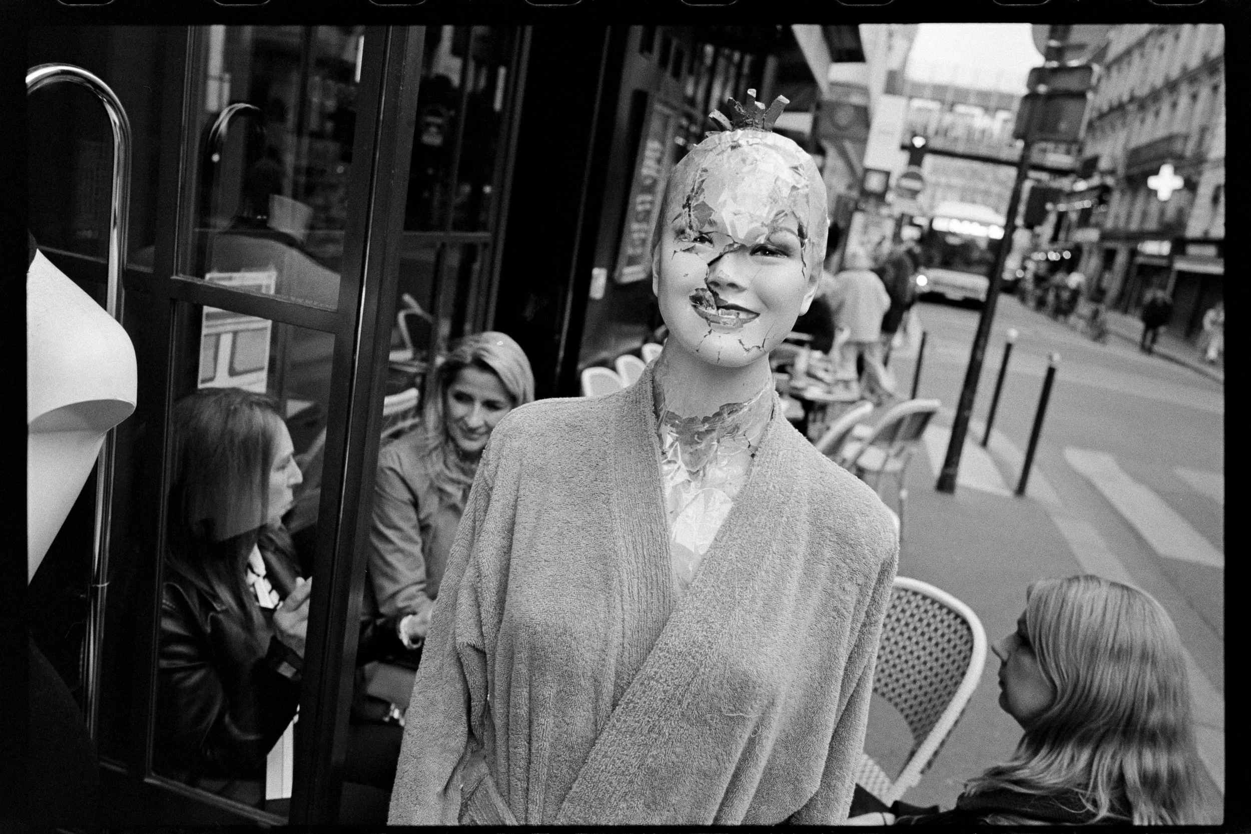 Une statue ou un mannequin représentant une personne avec un visage fissuré ou endommagé, portant un vêtement léger, debout sur un trottoir devant un café ou un restaurant à Paris, avec des personnes assises à une table voisine. La scène est dans une