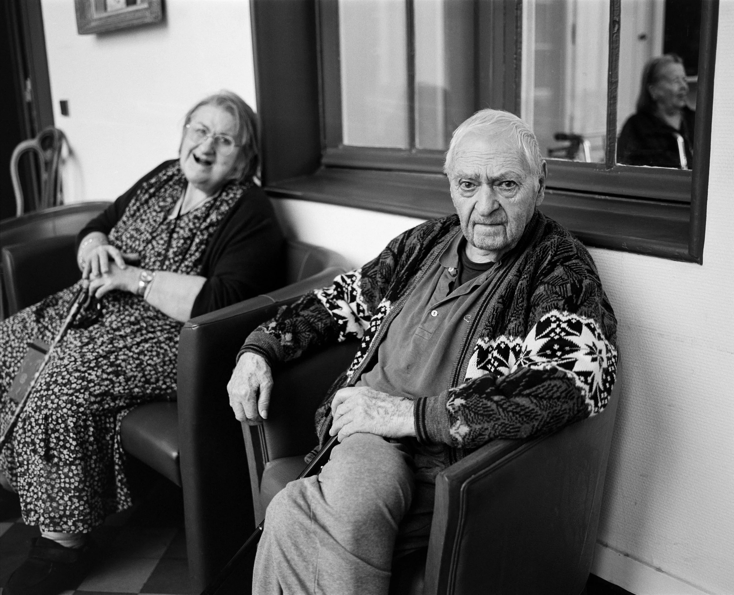 Deux personnes âgées assises dans une salle d'attente, une femme souriante portant un manteau noir et une robe à motifs, un homme sérieux portant un cardigan et une chemise, chacun avec une canne.