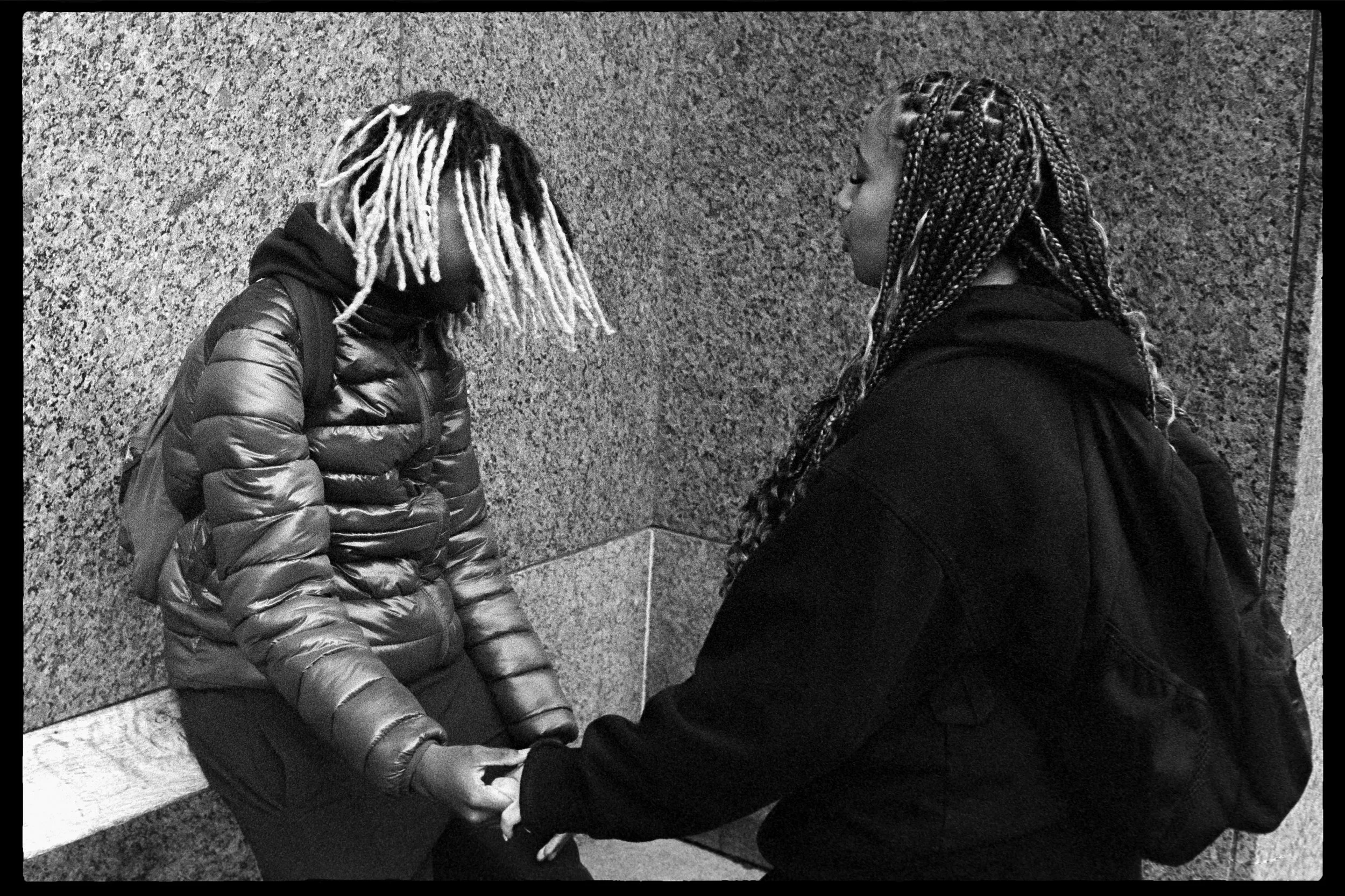 Deux femmes noire, dont l'une a des cheveux en dreadlocks blanches, se tiennent la main dans un endroit urbain, contre un mur en pierre, dans une posture sérieuse.