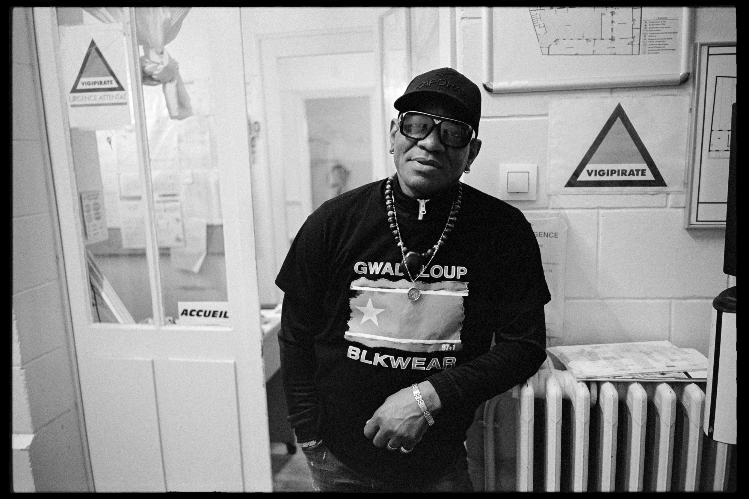 Homme portant un t-shirt noir avec un drapeau et des mots en anglais, portant des bijoux, des lunettes de soleil, et un bonnet, debout dans une pièce en intérieur.