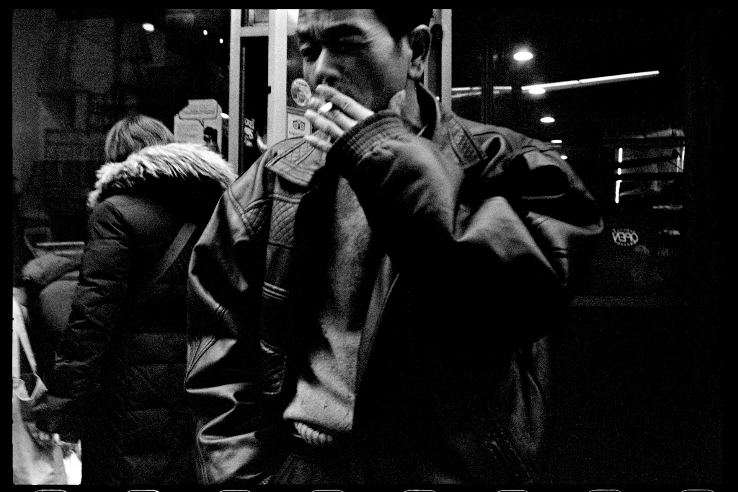 Un homme portant un manteau en cuir noir, en train de fumer devant une fenêtre, dans un environnement urbain nocturne.