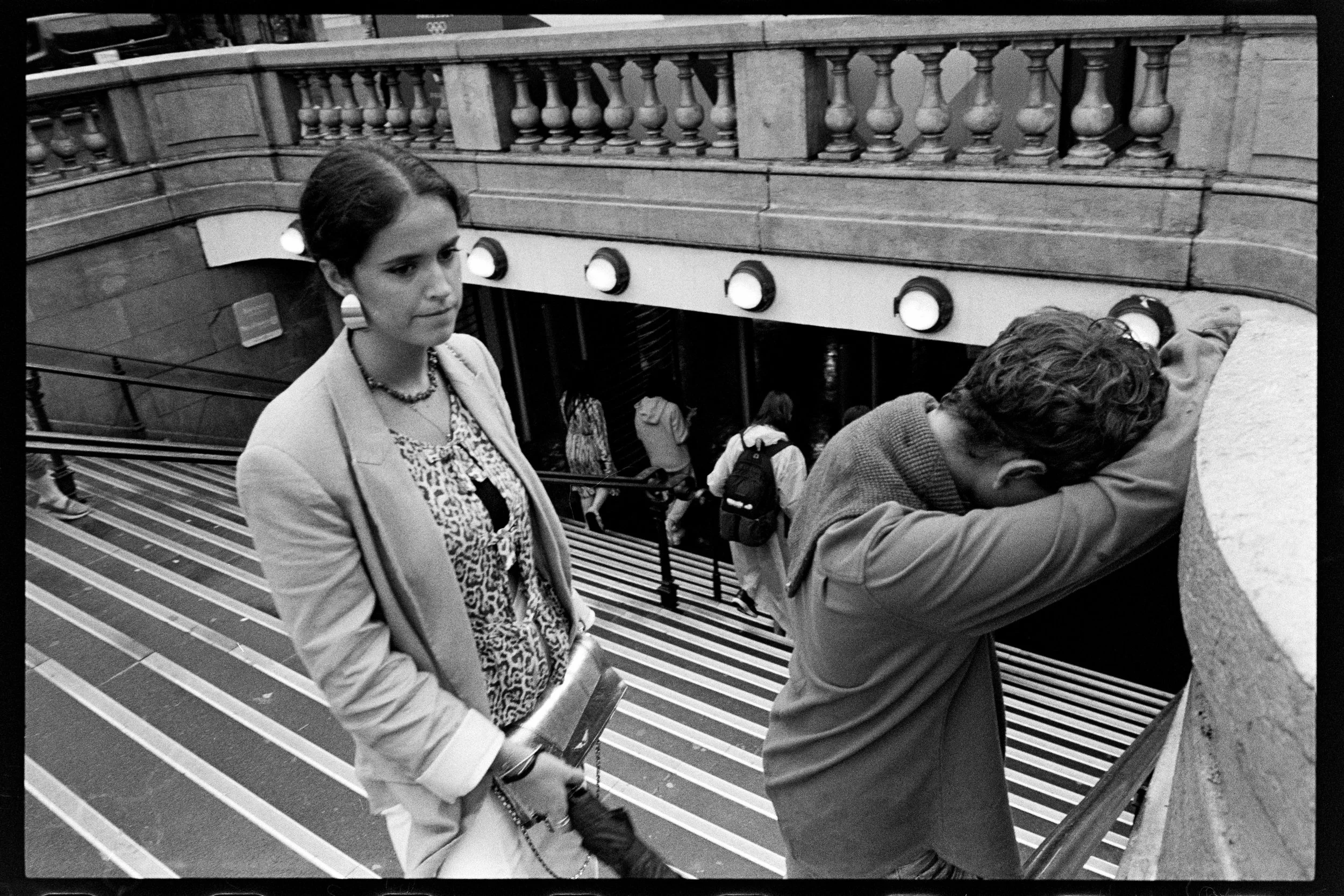 Une jeune femme en manteau clair et vêtements à motifs, une jeune homme penché contre une rambarde, dans un bâtiment avec un escalier intérieur en marbre et des lumières rondes, des personnes avec des sacs à dos passant en arrière-plan.