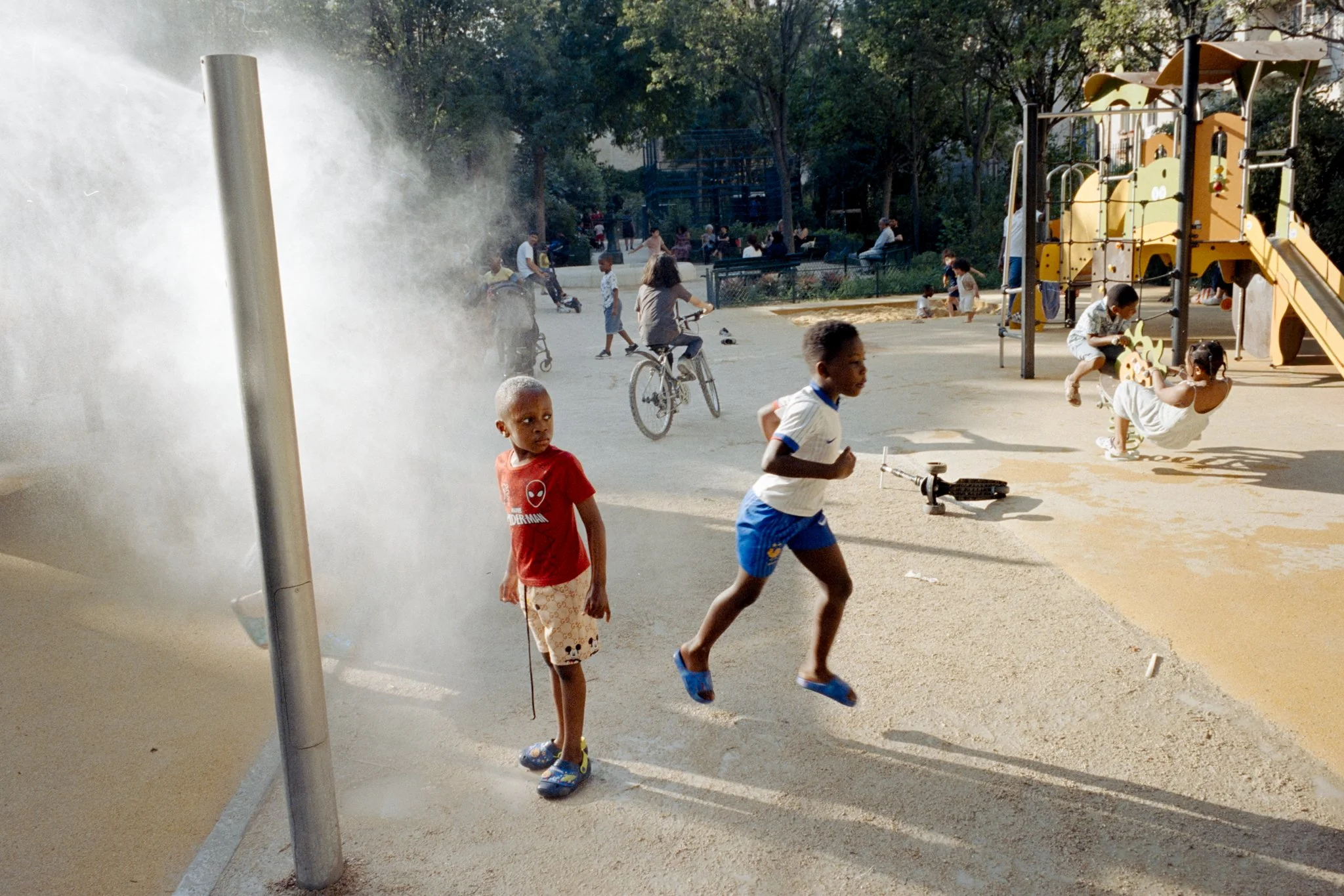 Enfants jouent et courent dans un parc urbain. Deux garçons en premier plan, un à côté de la buse d'incendie et l'autre qui court, un garçon qui fait du vélo à l'arrière, et des enfants sur une structure de jeu.