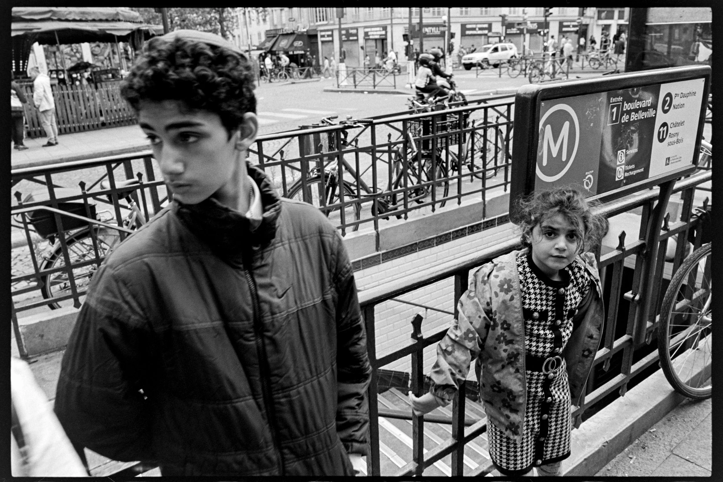Deux enfants se tiennent à l'entrée d'une station de métro à Paris, l'un garçon avec une veste foncée et une fille avec une robe à carreaux et un manteau floral, dans une rue animée.